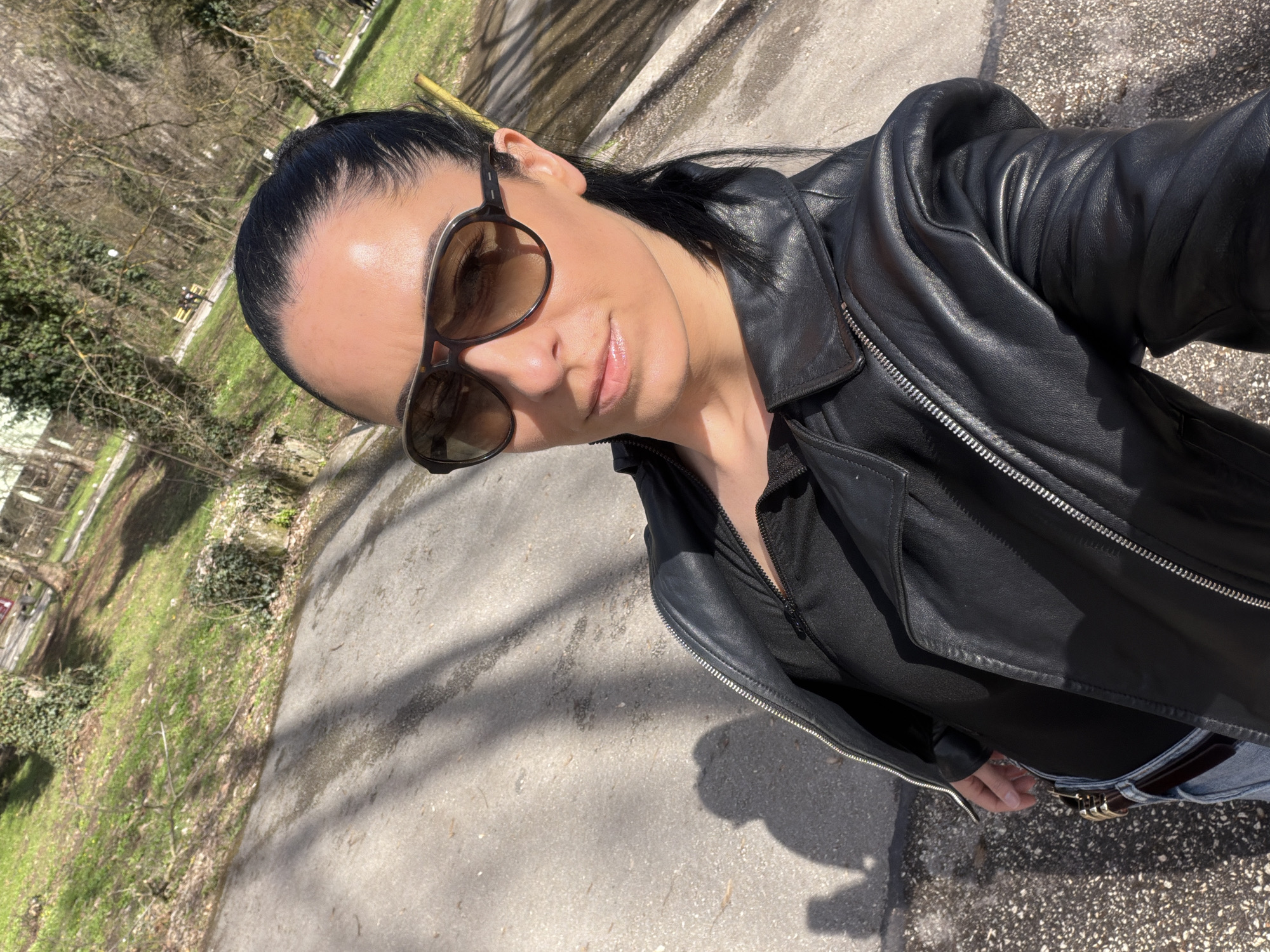 A solitary individual, appearing female, is depicted in a close-up selfie, standing outdoors in a paved area within Pleven, Bulgaria. The subject wears large aviator-style sunglasses with brown-tinted lenses, a black leather jacket unzipped to reveal a dark garment underneath, and light-colored jeans secured by a dark brown belt with a golden buckle. Her dark hair is pulled back, and her face is directly angled towards the camera. Strong sunlight from the right casts distinct shadows on the asphalt-like ground in the foreground. The background consists of a sloping, grassy terrain with deciduous trees, some covered in ivy, indicating a park or green space. A distant yellow park bench and faint structural elements are visible amongst the trees.