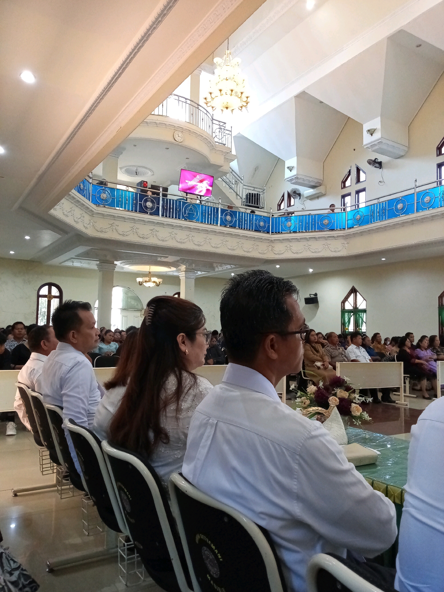The image depicts an indoor gathering, likely a religious service or event, taking place in the Grand Ballroom of the Hotel Sahid Manado, located in Bitung, Indonesia. The hall features a multi-tiered architectural design with a prominent second-level balcony. The main floor is filled with attendees seated in rows of chairs, facing a central area. The attendees, predominantly adults, are dressed formally, with many men wearing white collared shirts. In the foreground, a man with glasses and a white shirt is seen from the back, facing away from the camera. A woman with her hair in a bun is seated next to him. The background reveals the hall's ornate ceiling, a chandelier, and a large screen displaying pink graphics. The balcony area shows people standing and looking down. The overall ambiance is one of a formal assembly within a well-decorated indoor venue.
