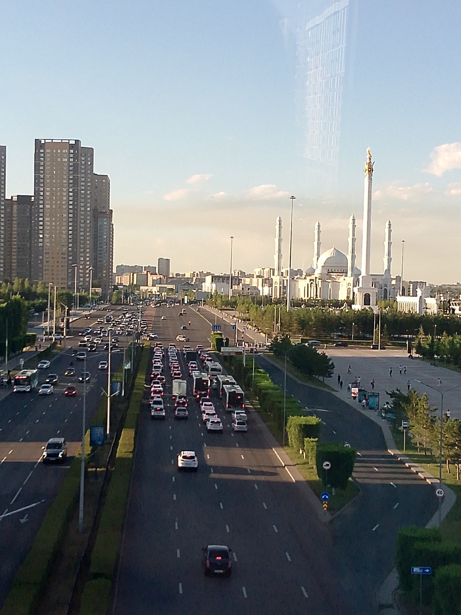 This is a high-angle shot of a wide, multi-lane road with moderate traffic. The scene is an urban environment with tall buildings on the left and a large, white mosque with several minarets on the right. A prominent golden spire atop a tall, slender monument stands near the mosque. The road is lined with trees and lampposts, with a central median. Cars and buses are visible in various lanes, indicating movement. People are seen walking on a pedestrian area adjacent to the road on the right side, and a few individuals are visible on the sidewalk on the left. The sky is clear with a few wispy clouds, suggesting it is daytime, likely late afternoon or early evening given the warm, soft light. The overall atmosphere is calm and busy. No discernible text is visible in the image.
