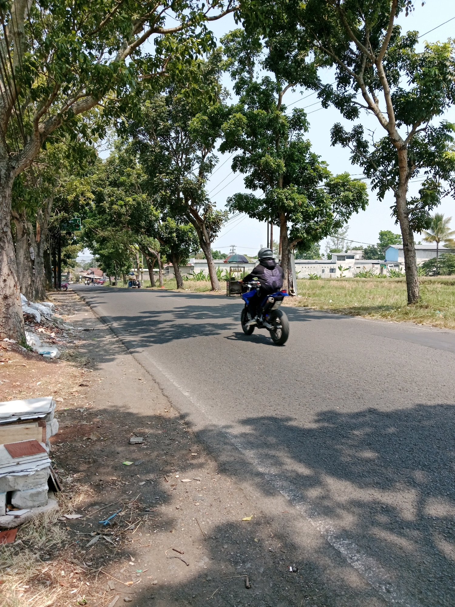 A motorcycle rider wearing a helmet and a dark jacket is riding on a paved road in Karangtengah, Indonesia. The rider is moving away from the camera, slightly to the right of the frame. The road is lined with trees on both sides, casting shadows on the asphalt. The background shows dry grass, some buildings, and utility poles. In the foreground, on the left side of the road, there are piles of white plastic bags and some construction materials. The sky is clear and blue, indicating a sunny day. Several other motorcycles are visible in the distance on the road.
