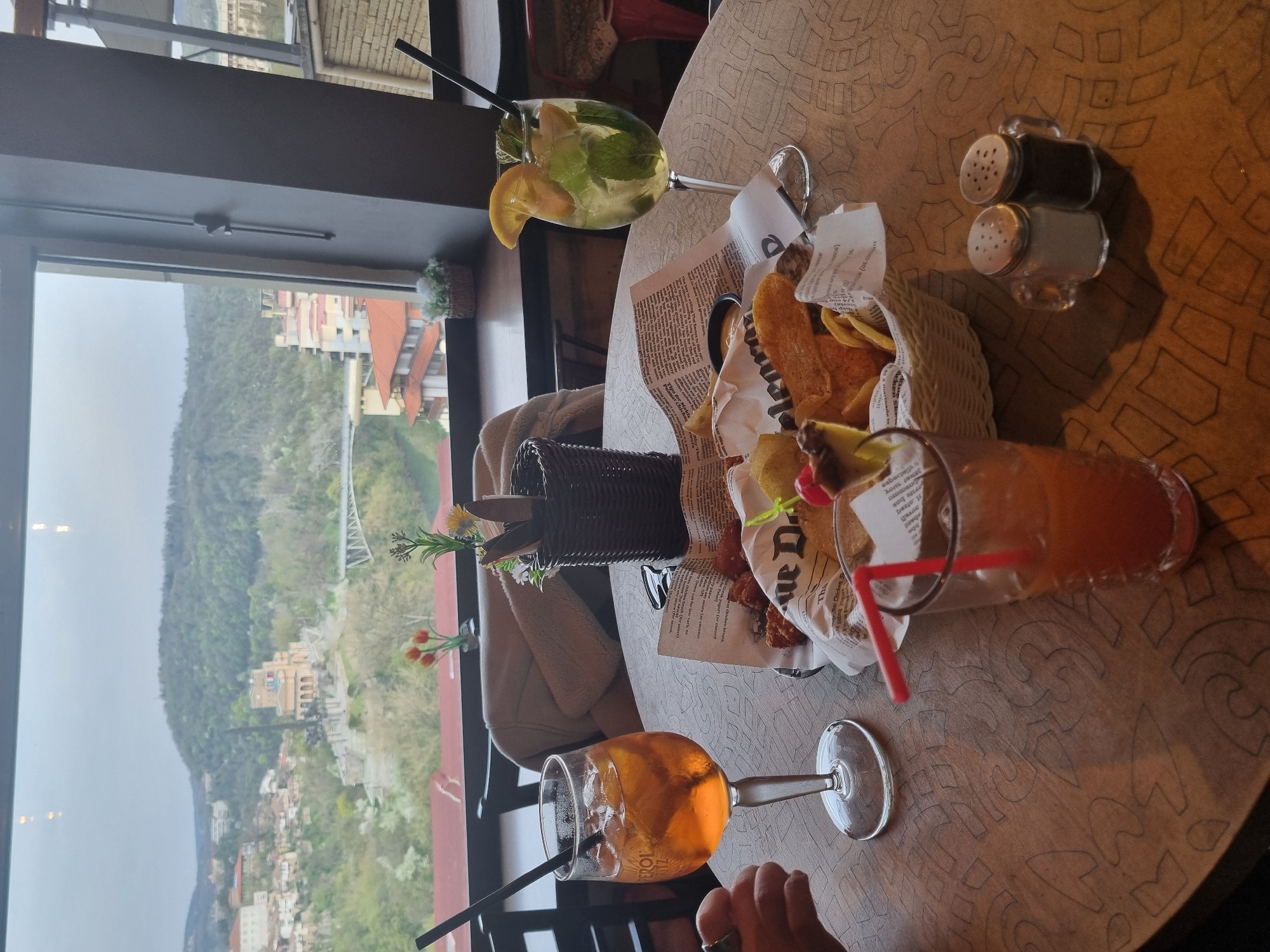 The image shows a table in a restaurant in Veliko Tarnovo, Bulgaria, with a view of the Yantra River valley and the Stambolov Bridge. On the table, there are three cocktails: an Aperol Spritz, a Mojito, and another orange-colored cocktail. There's also a basket of fried snacks, a small decorative plant in a wicker pot, and a salt and pepper shaker. A person's hand is visible at the bottom of the frame. The table has a patterned wooden surface. Through the window, a cityscape stretches across the river valley, with the Stambolov Bridge visible in the distance. The weather appears to be overcast.