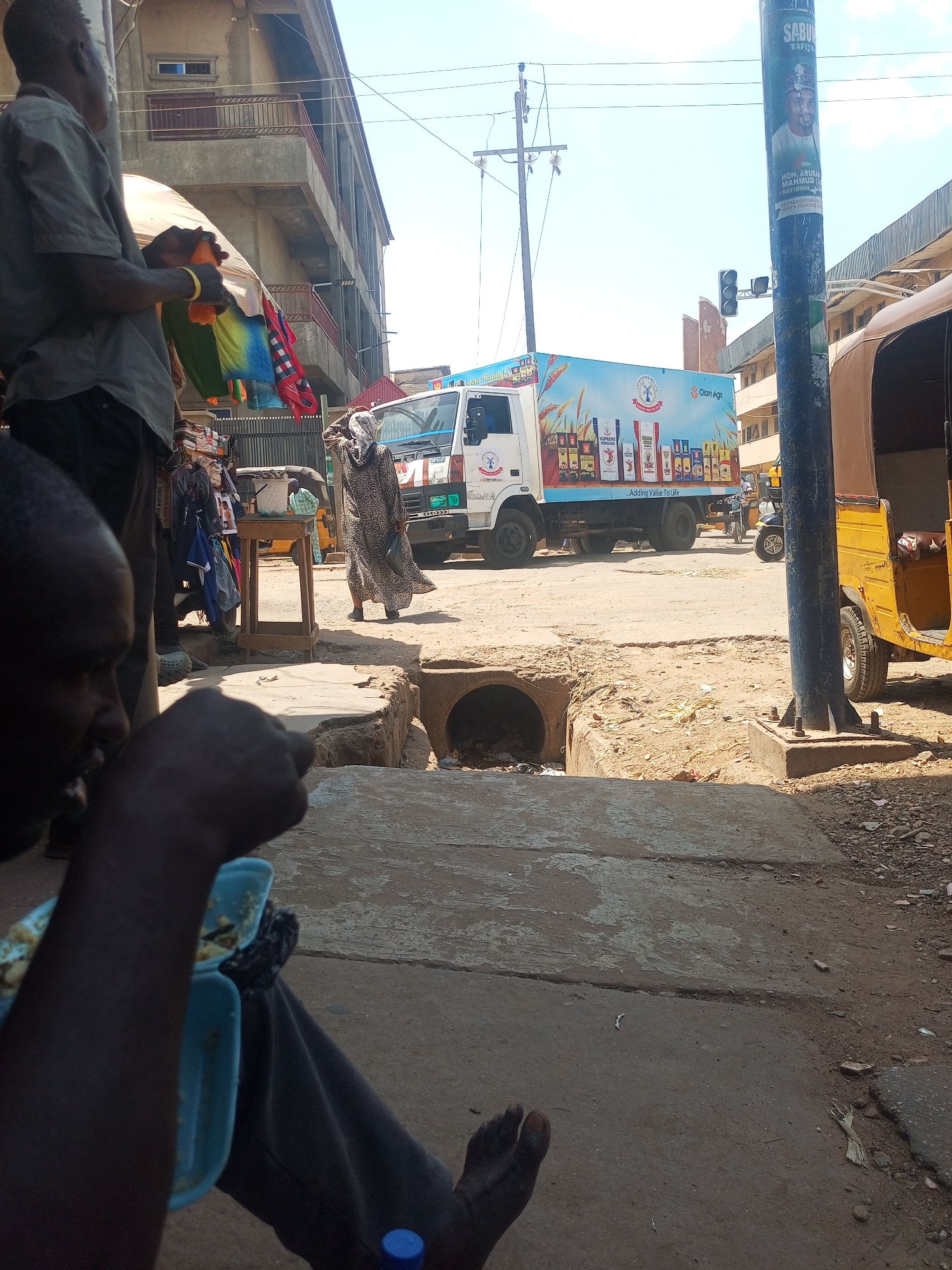 Here's a detailed description of the scene:
**Location:** Maiburiji, Nigeria.
**Overview:** The image depicts a bustling street scene. The scene is sunlit, with a mixture of dirt and paved surfaces. Several individuals are present, along with vehicles and commercial elements.
**People:**
* A man is prominently featured in the foreground eating food from a blue container.
* Another man stands nearby, holding colorful items (possibly clothing or fabric).
* A woman wearing a patterned dress and head covering is walking across the street.
* Other people are visible in the background, though less distinctly.
**Objects:**
* A white commercial truck with branding for "Olam Agri,".
* A yellow three-wheeled vehicle (likely an auto-rickshaw or tuk-tuk).
* A utility pole with a poster of a man and the word "SABU".
* A drainage ditch or culvert.
* Various stalls and makeshift stands.
**Actions and Interactions:**
* The man in the foreground is eating.
* The woman is walking across the street.
* The men stand near what appears to be merchandise, potentially engaged in commerce.
* A food vendor on the other side of the street.
**Environment:**
* Buildings are visible in the background.
* The street appears somewhat unpaved and dusty.
* The light suggests a sunny day.
* Traffic light.