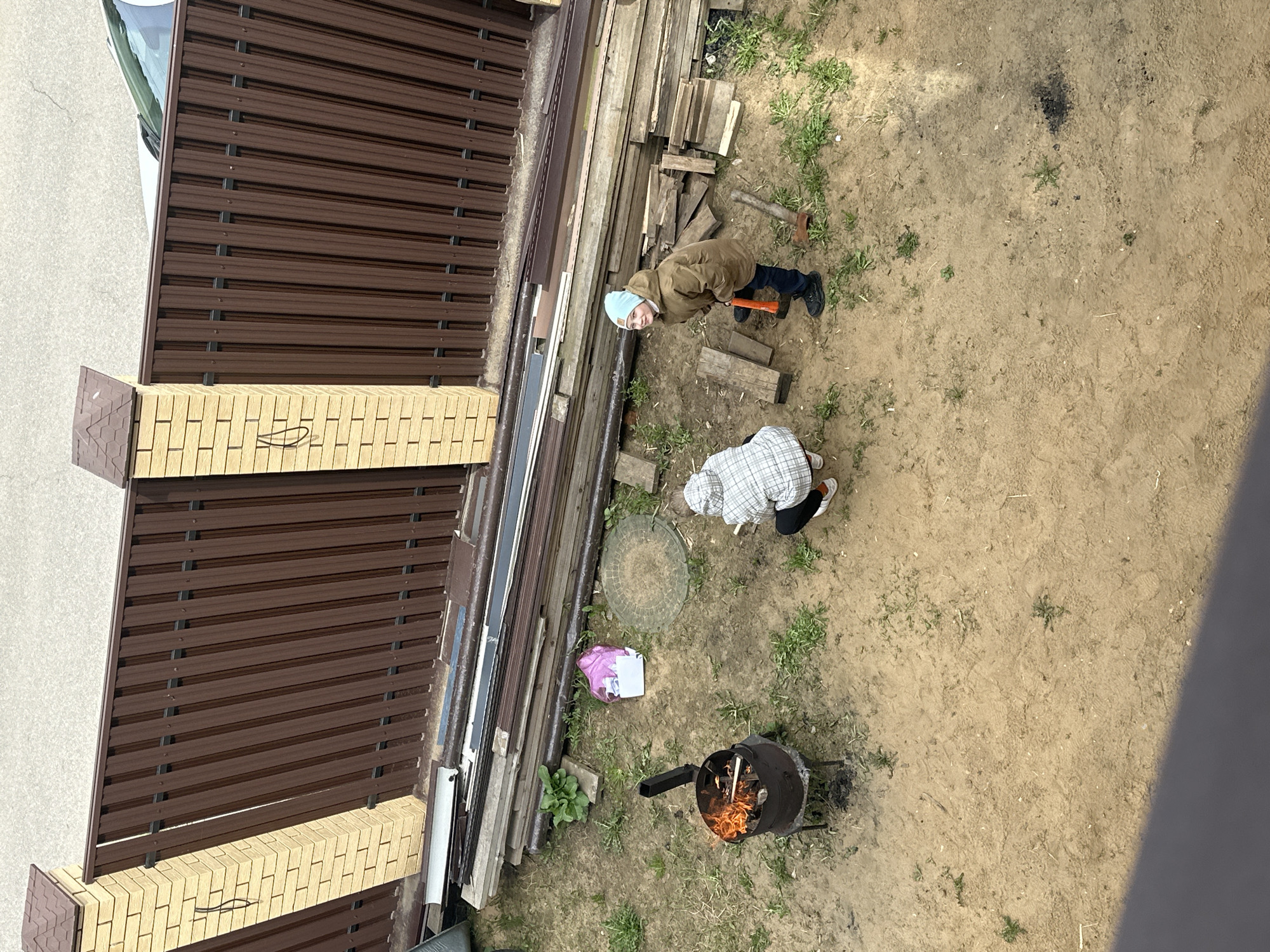 The image shows an overhead view of a backyard area, presumably in слобода Старая, Russia. A section of brown metal fence with a brick base runs along the left edge of the frame. Adjacent to the fence are stacks of lumber and various building materials. Two people are visible: one wearing a brown jacket and a light blue head covering is standing and holding an orange-handled tool. The second person, wearing a white and gray patterned head covering, is bending over. A black metal grill or incinerator containing burning wood is positioned in the lower part of the frame. The ground is mostly dirt with patches of grass. A plastic bag and a round manhole cover are near the lumber pile.