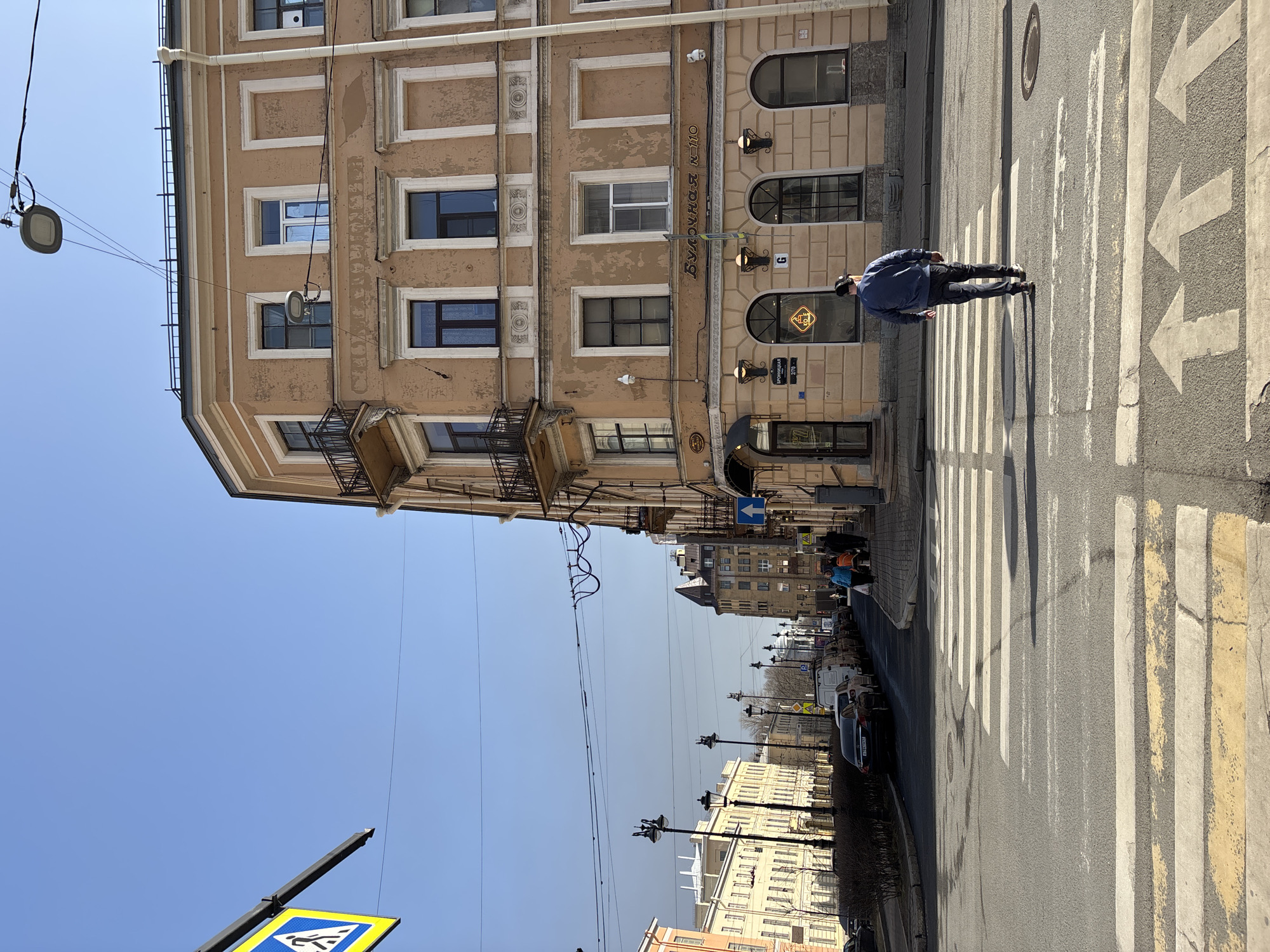 The image depicts an urban street scene at the intersection of Bronnitskaya Street and 1st Krasnoarmeyskaya Street in Saint Petersburg, Russia, under a clear blue sky. A light brown, five-story corner building with decorative window surrounds and multiple balconies dominates the right side, its facade showing signs of wear. The ground floor of this building features a commercial establishment identified as "Булочная №110" (Bakery No. 110) with arched windows and outdoor wall sconces, alongside a darker entrance at 27B Bronnitskaya Street.
In the foreground, a pedestrian crossing with white and yellow stripes extends across the asphalt road, accompanied by three forward-pointing white arrows painted on the road surface. A male figure, viewed from behind, wearing a blue jacket, dark pants, and a dark baseball cap, walks across this zebra crossing from left to right.
To the left of the crossing, a row of parked vehicles, including a dark sedan and several white vans, lines the street. Further down the street, numerous classical-style buildings with multiple windows are visible, along with multiple ornate streetlights. Overhead power lines and a pedestrian crossing sign on a pole are visible in the upper left. On the sidewalk to the right, several other individuals are walking further down the street. The scene is illuminated by strong sunlight, casting distinct shadows.