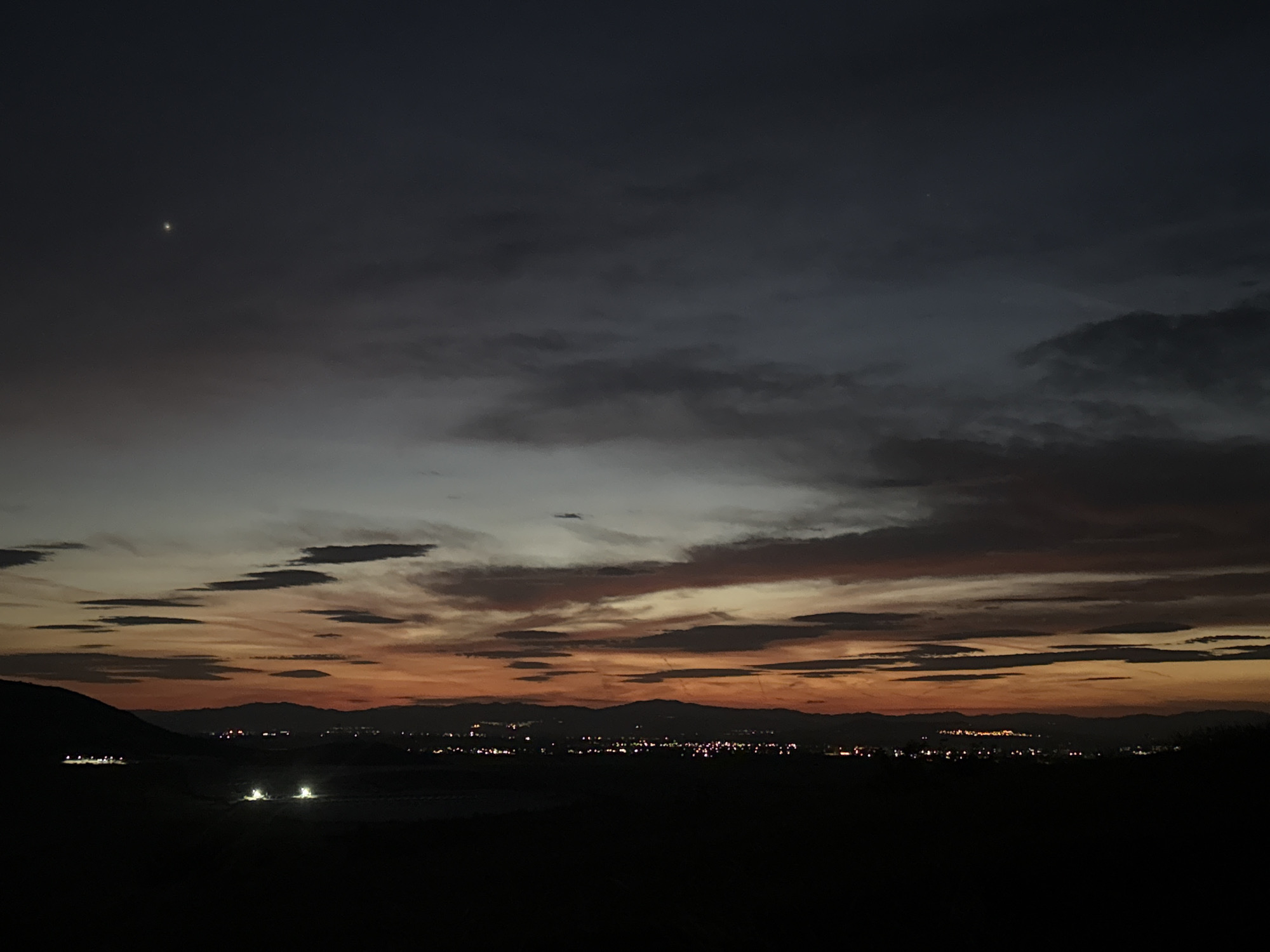 The image depicts a night or dusk landscape possibly near Sinitovo, Bulgaria. The sky is a gradient of colors, with a dark blue or black at the top transitioning to orange and red hues near the horizon. Several cloud formations are present. A bright point is visible in the upper left part of the sky, possibly a star or planet. On the horizon, there's a silhouette of distant hills or mountains. City lights are scattered along the horizon, suggesting a town or urban area. The foreground is dark and appears to be a field or open area. Several lights are visible in the near field, perhaps from buildings.