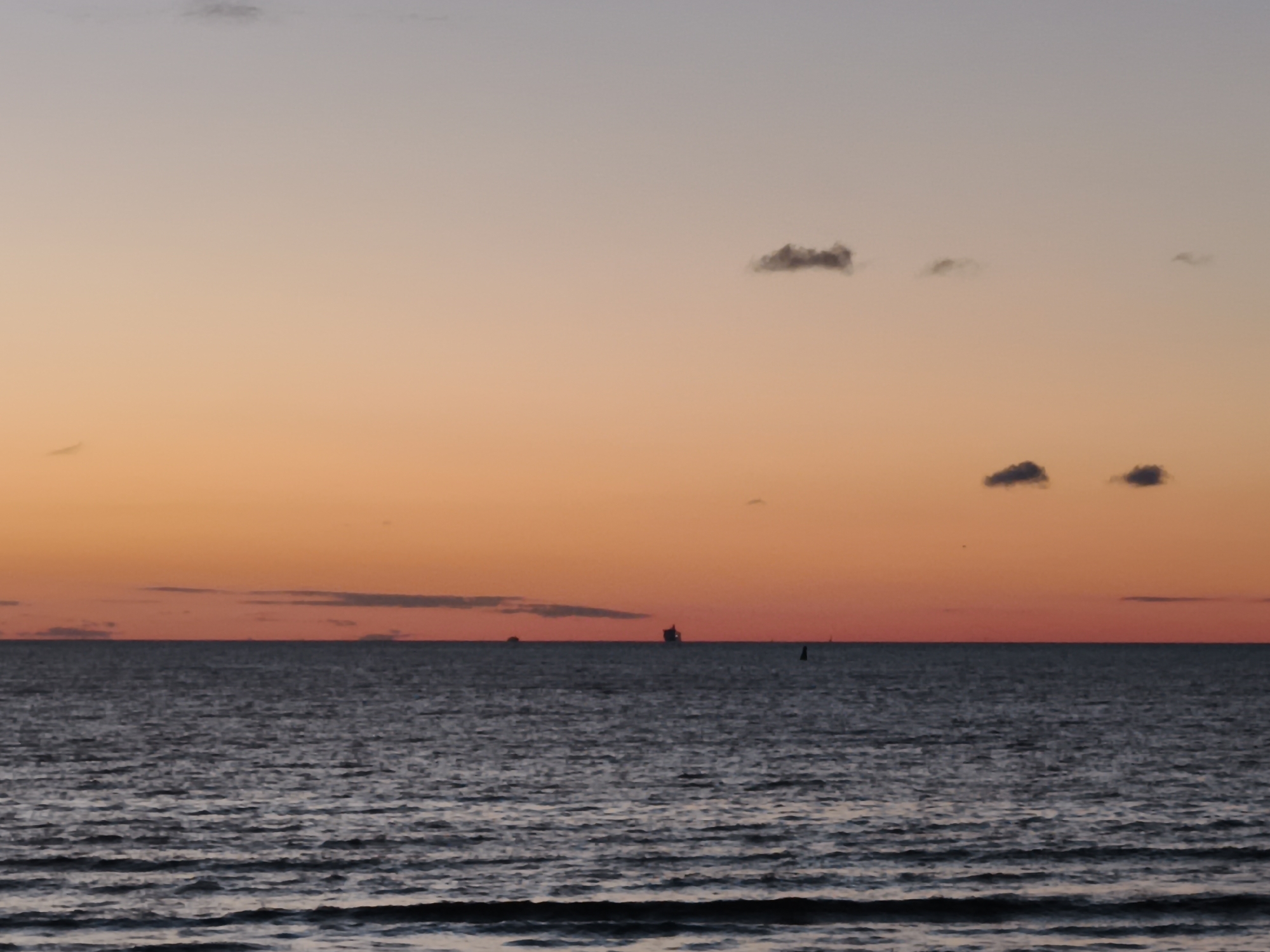 The image shows a serene seascape at sunset. The sky transitions from a pale blue at the top to a vibrant orange hue closer to the horizon. Several scattered clouds are visible in the sky. The sea is calm with gentle waves reflecting the colors of the sky. In the distance, several ships and a buoy are visible on the water. The scene suggests a tranquil evening by the coast of Tallinn, Estonia.