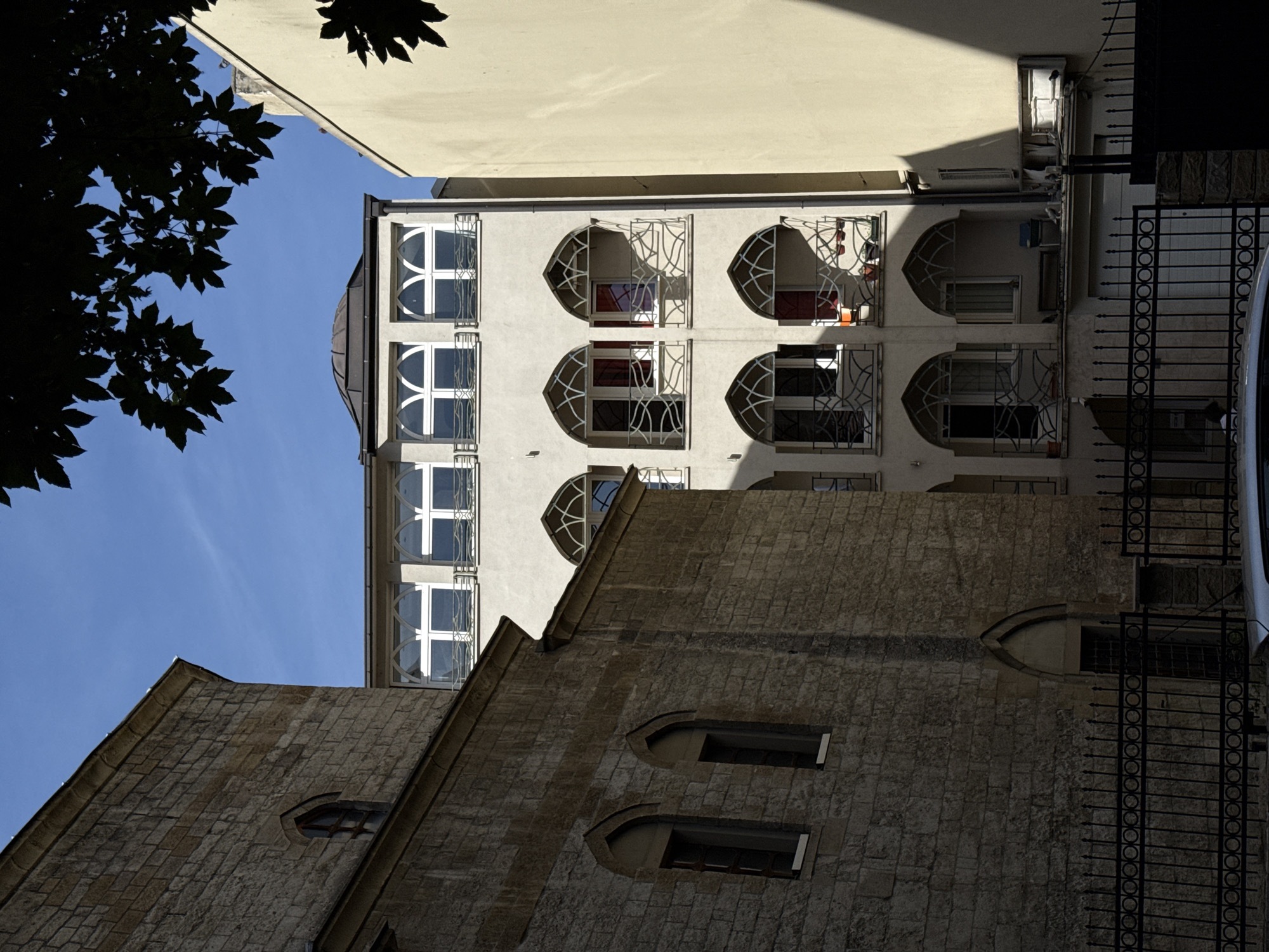 The image shows buildings in Belgrade, Serbia. In the foreground is a stone building with pointed arched windows. Above it is a taller white building with curved windows, some featuring ornamental metalwork. A black metal fence runs along the side of the buildings. The sky is visible in the background, along with the edge of another building. A tree branch with leaves is visible in the upper left corner.
