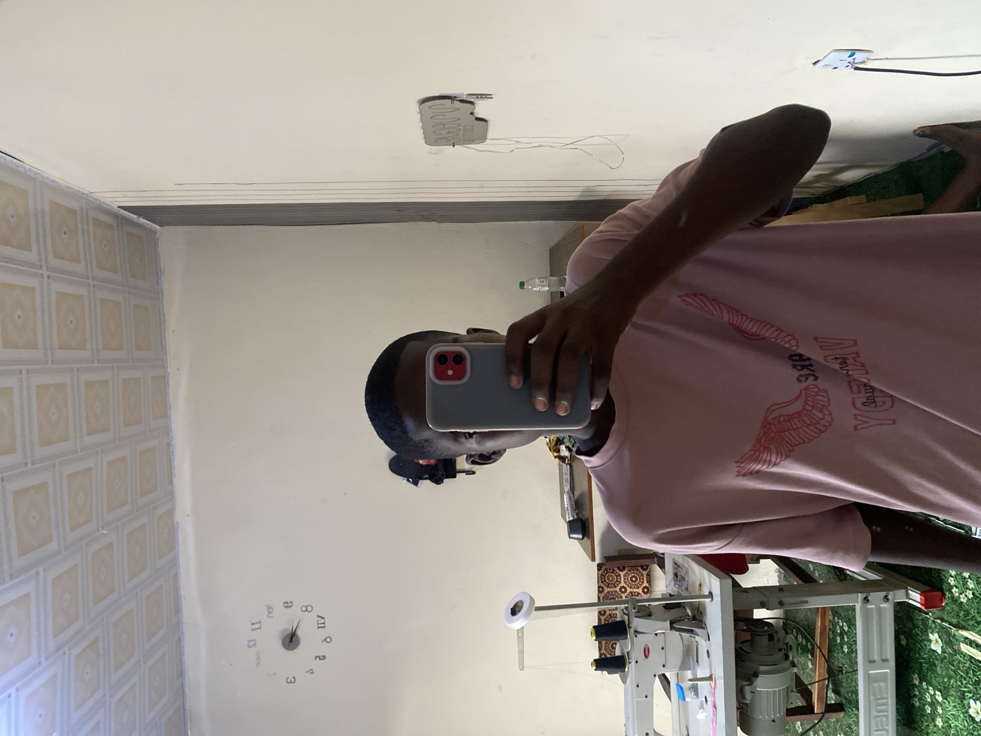 The image shows a young person taking a mirror selfie. The person is wearing a pink t-shirt with a graphic design of wings and text. They are holding a smartphone up to take the picture. In the background, there is a sewing machine with spools of thread, a clock on the wall, and a light-colored ceiling with a patterned tile design. There is also a wall outlet with a cable plugged in. The overall environment appears to be an indoor space, possibly a workshop or a bedroom. The location is stated to be in Jauro Yinu, Nigeria.