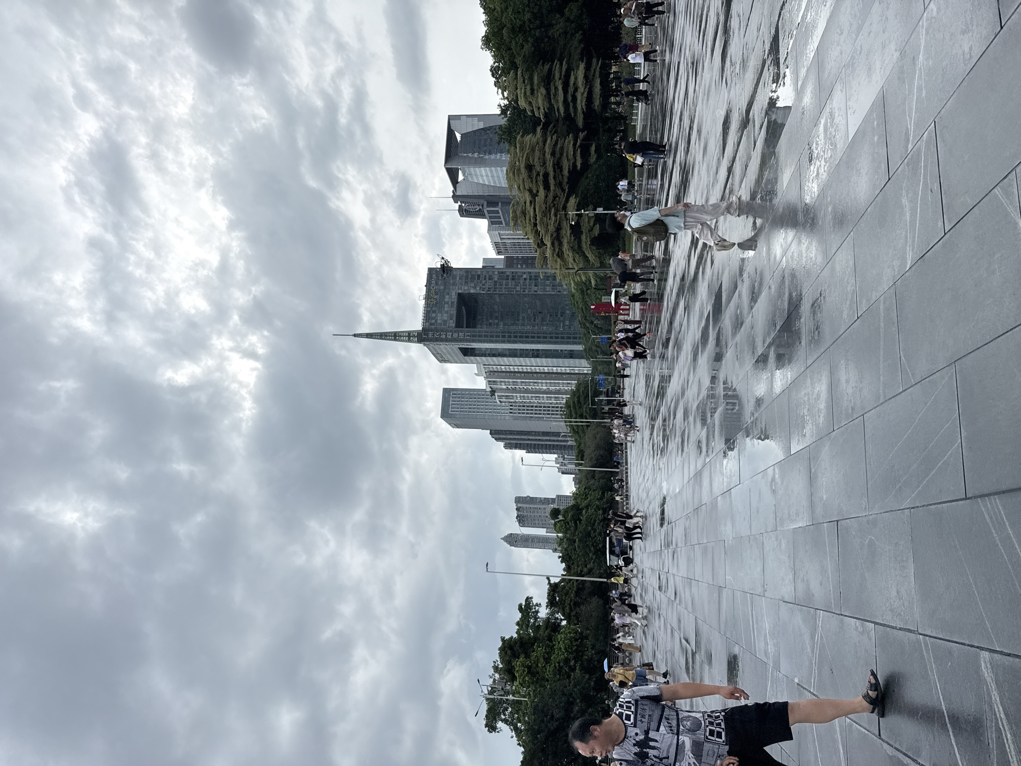 The image depicts a wide, wet, paved plaza in Tianhe District, China, under a heavily overcast sky. The plaza floor is made of large, dark gray tiles with white veining, reflecting the muted light and the figures of people walking across it. Numerous individuals are scattered across the plaza, some walking alone, others in small groups. In the background, several modern skyscrapers of varying heights and architectural styles dominate the skyline, interspersed with lush green trees. The overall atmosphere is somber due to the cloudy weather, and the wet ground suggests recent rain. The foreground features a man in a patterned t-shirt and dark shorts walking towards the left side of the frame. Further back, another person with a backpack walks away from the camera. The scene captures a public space in a bustling urban environment.