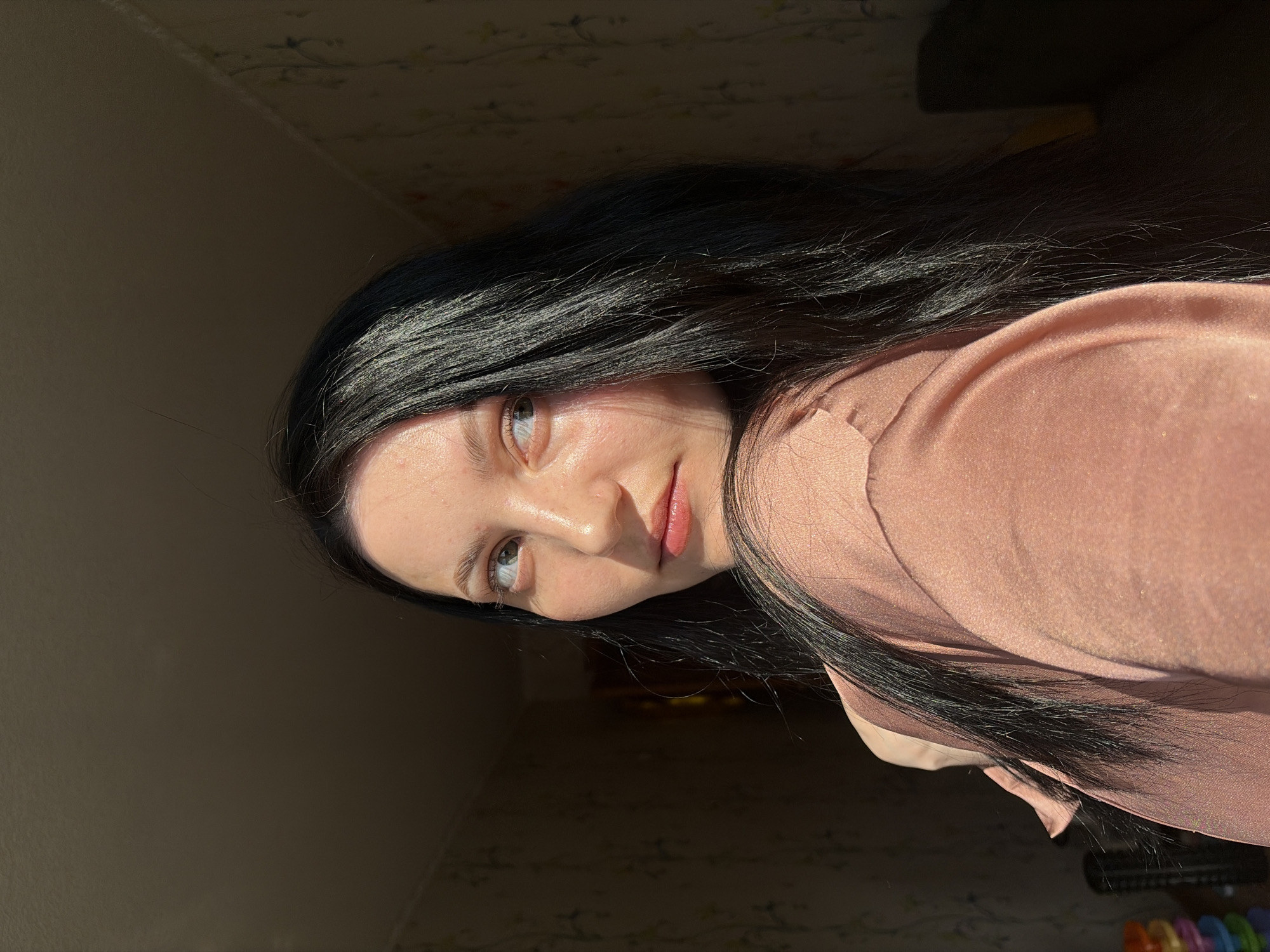A portrait of a young woman with long, dark hair is captured indoors. The light source illuminates the left side of her face, while the right side is cast in shadow. She has fair skin, light-colored eyes, and pink lips. She is wearing a pink satin top. The background consists of a beige wall with subtle patterns. The location is in a room, potentially in the city of Chaikovsky, Russia.