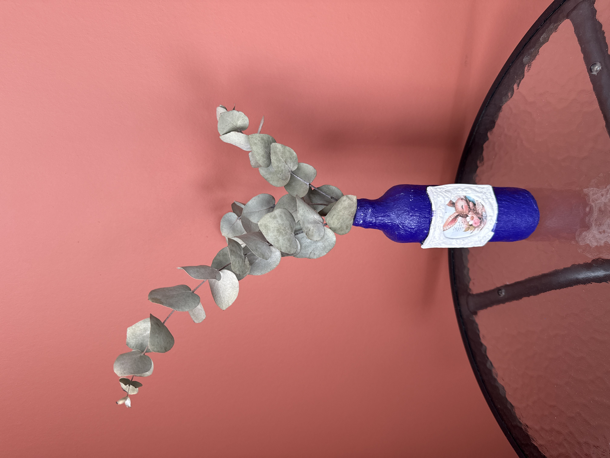 A blue, decorated bottle lies horizontally on a glass-topped table with a metal frame. The bottle contains sprigs of eucalyptus. The background is a plain, coral-colored wall. The location cannot be determined from the image.