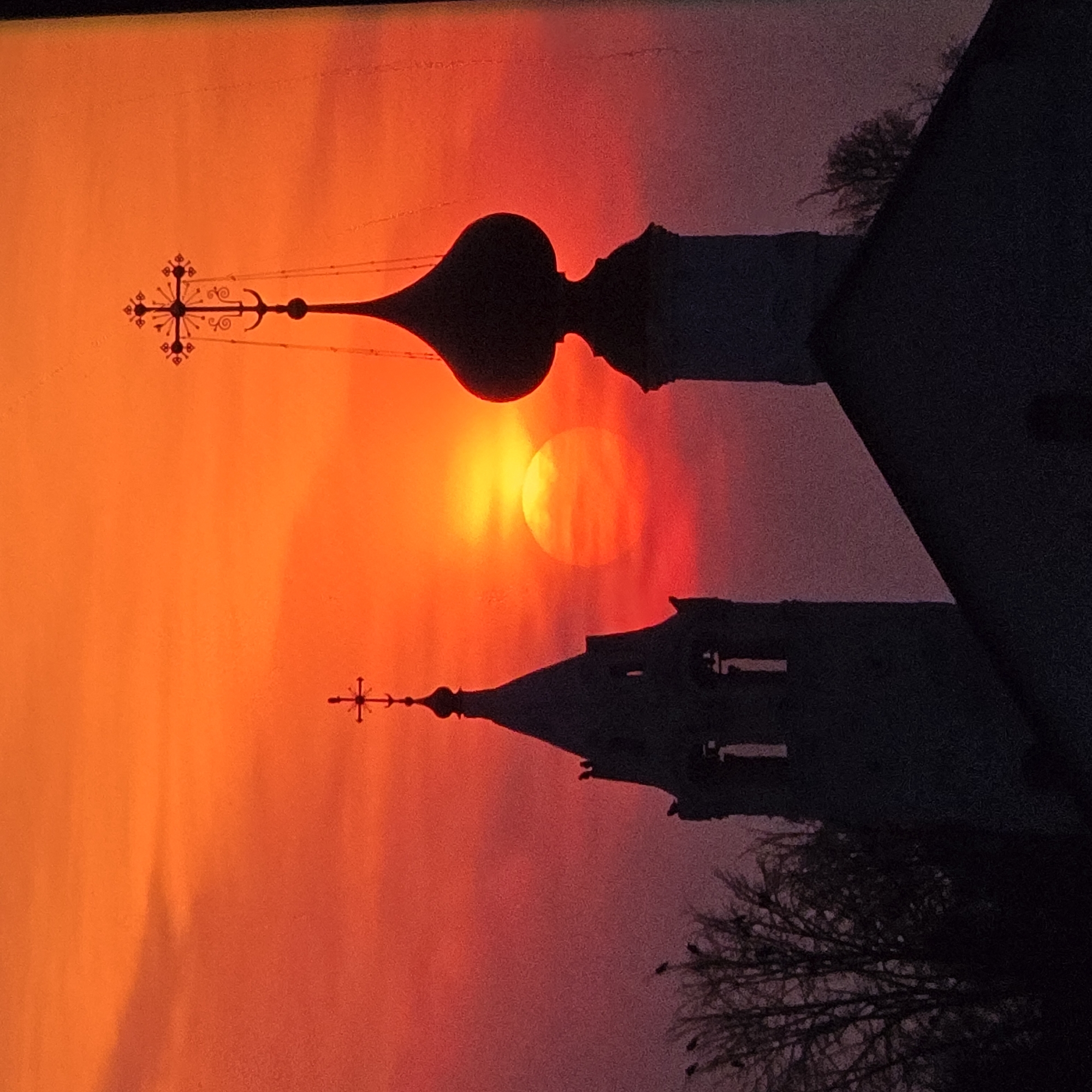 The image captures a silhouetted view of two church bell towers and a domed roof against a vibrant sunset sky, likely in Vladimir, Russia, given the architectural style which resembles onion domes typical of Russian Orthodox churches. The sky is dominated by intense shades of orange and red, with the sun appearing as a bright, partially obscured orb in the center-left of the frame.
On the left, a taller, more slender bell tower is visible, topped with a cross. Its structure features arched openings and decorative elements. Birds are perched on the tower and on the bare branches of a nearby tree, also silhouetted against the sky.
To the right, a second, wider bell tower is depicted, topped with a distinctive onion dome and a cross. The silhouette of a building with a sloped roof, possibly part of the church complex, extends from the base of this tower towards the foreground.
The overall scene is one of stark contrasts between the dark silhouettes of the architecture and the brilliant, fiery hues of the sunset. The image is taken from a ground-level perspective, looking upwards towards the sky and the prominent religious structures. There are no visible people in the frame. The environment appears to be an outdoor setting, likely within the grounds of a church or monastery.