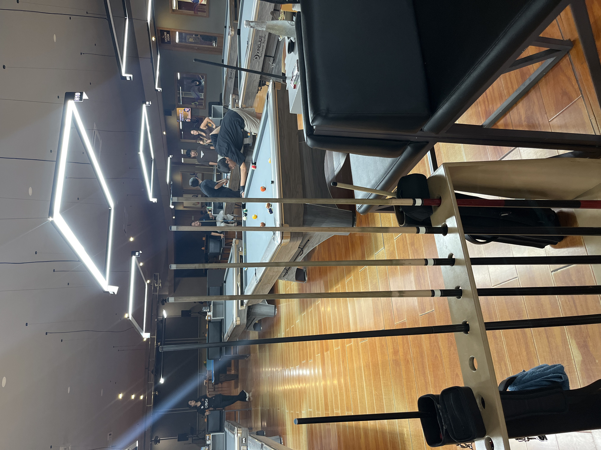 This media file depicts an indoor pool hall with several pool tables. The scene is illuminated by modern, angular fluorescent lights. In the foreground, a rack of pool cues is visible, some with colorful tips. The floor is made of polished wood.
Several people are present in the scene. Some are standing around the pool tables, possibly waiting to play or observing a game. One individual, wearing a black shirt with "ORCA" printed on it, is standing near a pool table. Another person in a black shirt is bent over a pool table, seemingly playing or setting up a shot. Further back, more individuals are seen, some seated and others standing.
The pool tables themselves appear well-maintained, with green felt surfaces and colorful billiard balls. The overall atmosphere suggests a casual social gathering or a recreational activity. The presence of pool tables and cues indicates a setting dedicated to playing pool. The style of the lighting and the polished wooden floor suggest a contemporary and somewhat upscale establishment. No specific time of day or weather conditions are discernible from the indoor setting. There is text visible on some of the pool cues and on the shirts of some individuals.