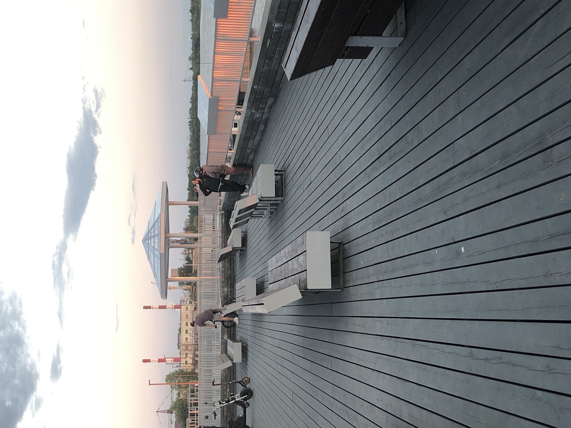 The image depicts an outdoor scene with a wooden deck leading into the distance. Two people are visible on the deck, one standing and looking out, the other seated on a bench. The setting appears to be a park or recreational area, with benches and some architectural structures in the background, including a glass-covered pavilion. The sky is overcast, suggesting cloudy weather. The time of day appears to be late afternoon or early evening, as indicated by the soft lighting. In the distance, there are industrial structures, including what appear to be smokestacks, and a body of water. The overall mood is peaceful and serene.