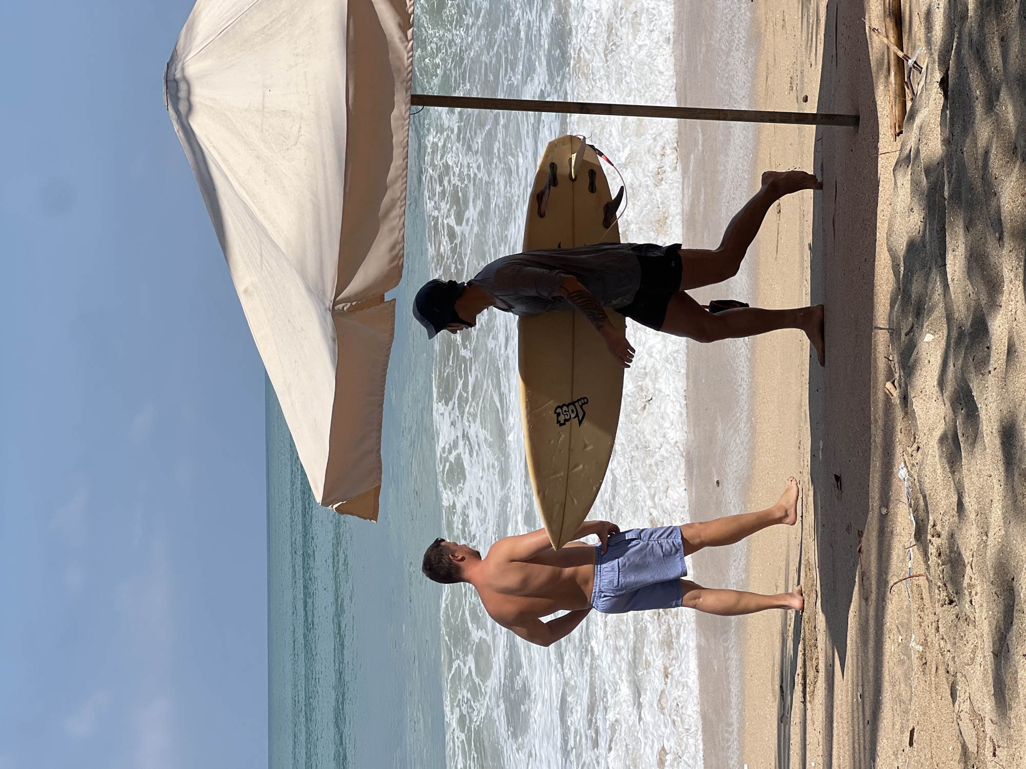 Two men are on a beach in Kuta, Indonesia. One man, facing away from the camera, is standing with his hands on his hips, looking out at the ocean. He is wearing blue and white striped swim trunks. The other man, wearing a dark helmet, grey t-shirt, and black shorts, is walking to the right, carrying a surfboard. The surfboard is yellow and has fins. A large, light-colored beach umbrella shades the two men. The ocean has waves, and the sky is clear and blue. The sand on the beach is light brown and has some debris.