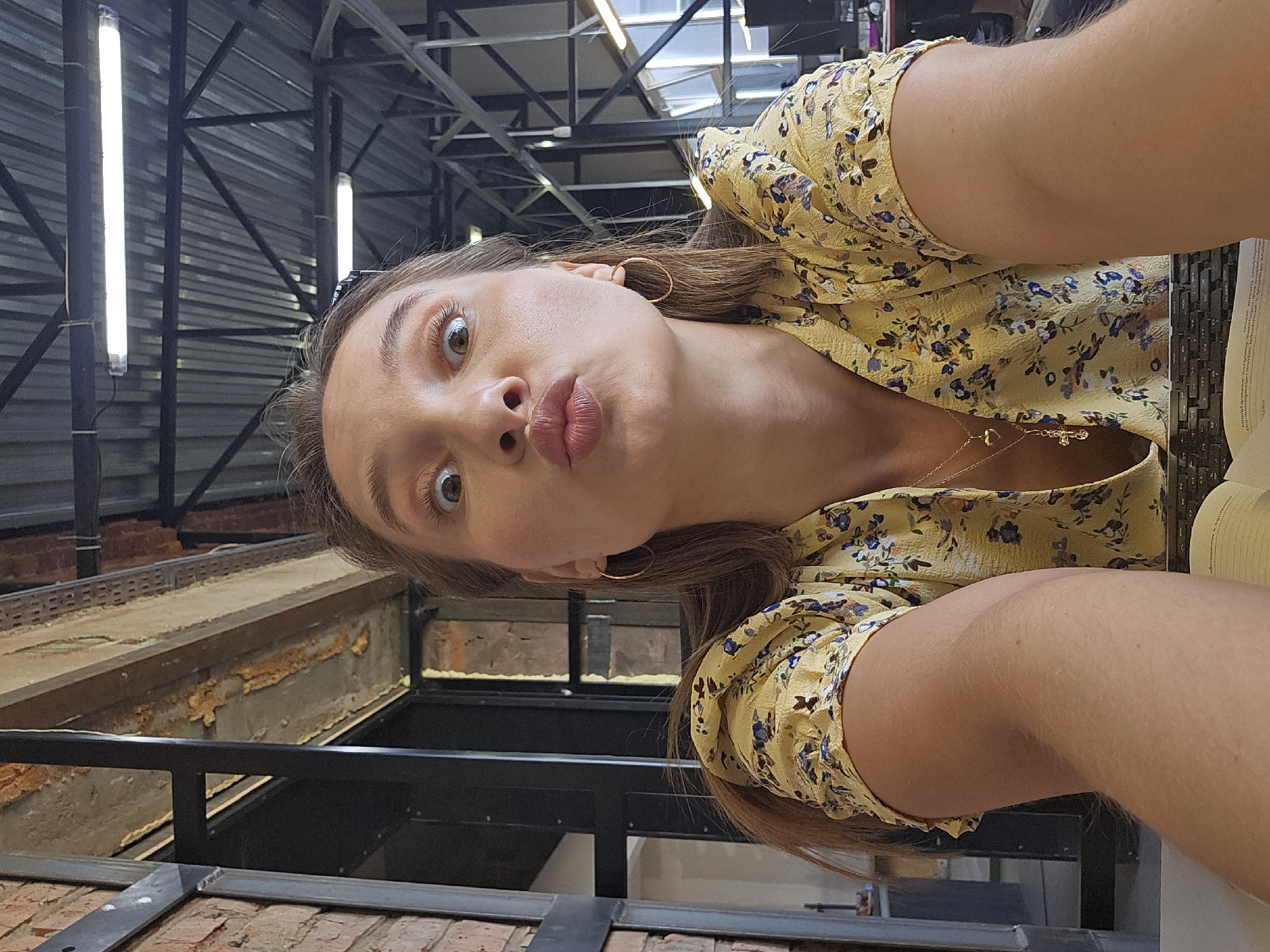 A woman with light skin, brown hair, and brown eyes is taking a selfie. She is wearing a yellow floral print top, gold hoop earrings, and a gold necklace. Her lips are pursed into a pucker. She appears to be in a building with exposed brick, metal beams, and fluorescent lighting. The exact location in Chaikovsky, Russia, is indeterminable from the image.