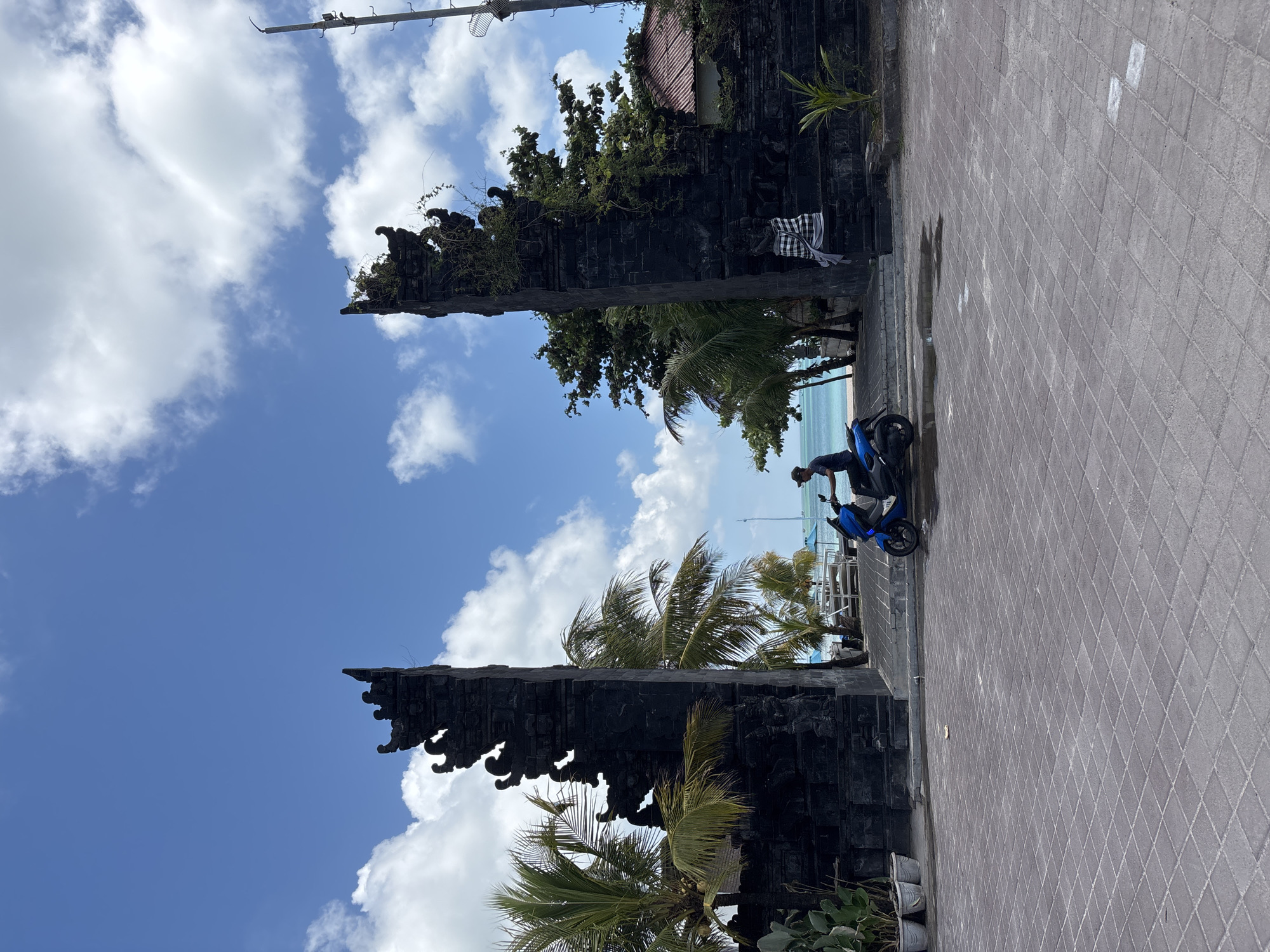 Captured in Kuta, Indonesia, the image shows a partially obscured blue sky with scattered clouds framed by two ornate, dark stone gate structures. Palm trees interspersed between the gates and the foreground add to the tropical atmosphere. A man is situated near a blue scooter parked on a paved walkway, which stretches into the frame with a diamond-shaped pattern. The environment suggests an outdoor setting, likely a public area near a beach or park, with the presence of both natural elements and human-made structures.
