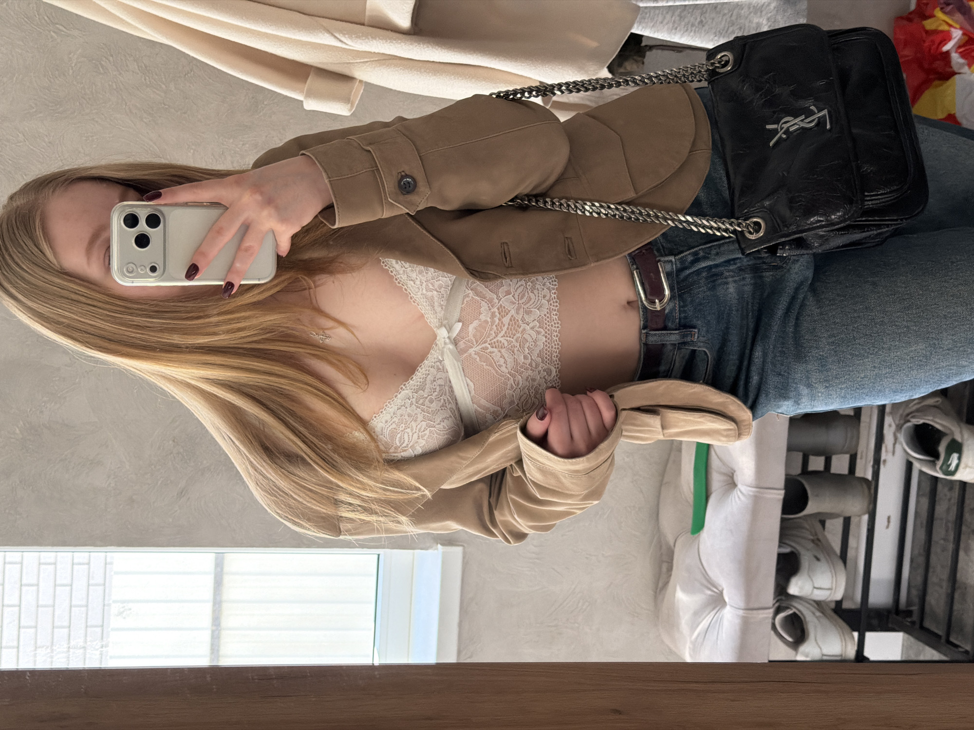 A blond woman is taking a selfie in a mirror. She's wearing a brown jacket over a white lace top, and blue jeans. A black YSL bag hangs from her shoulder. In the background, there is a white coat and a shoe rack with white sneakers. The reflection is likely in an apartment or house. The location is unknown, and the query for Rossiyskiy, Russia, is irrelevant to the contents of the image.