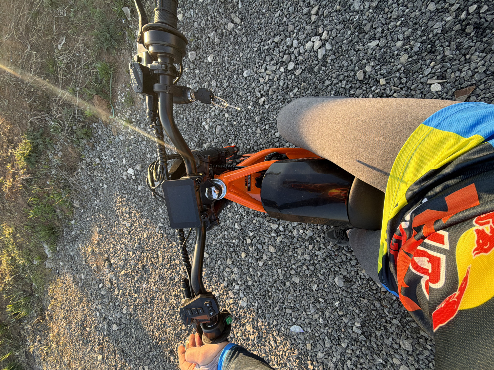 A high-angle shot shows the handlebars and part of the frame of an orange and black electric bicycle or motorcycle. A person wearing gray pants and a brightly colored (blue, yellow, orange, black, white) jersey is astride the vehicle. The ground surface is composed of small, gray gravel. The location is the city of Азьмушкино, Russia.