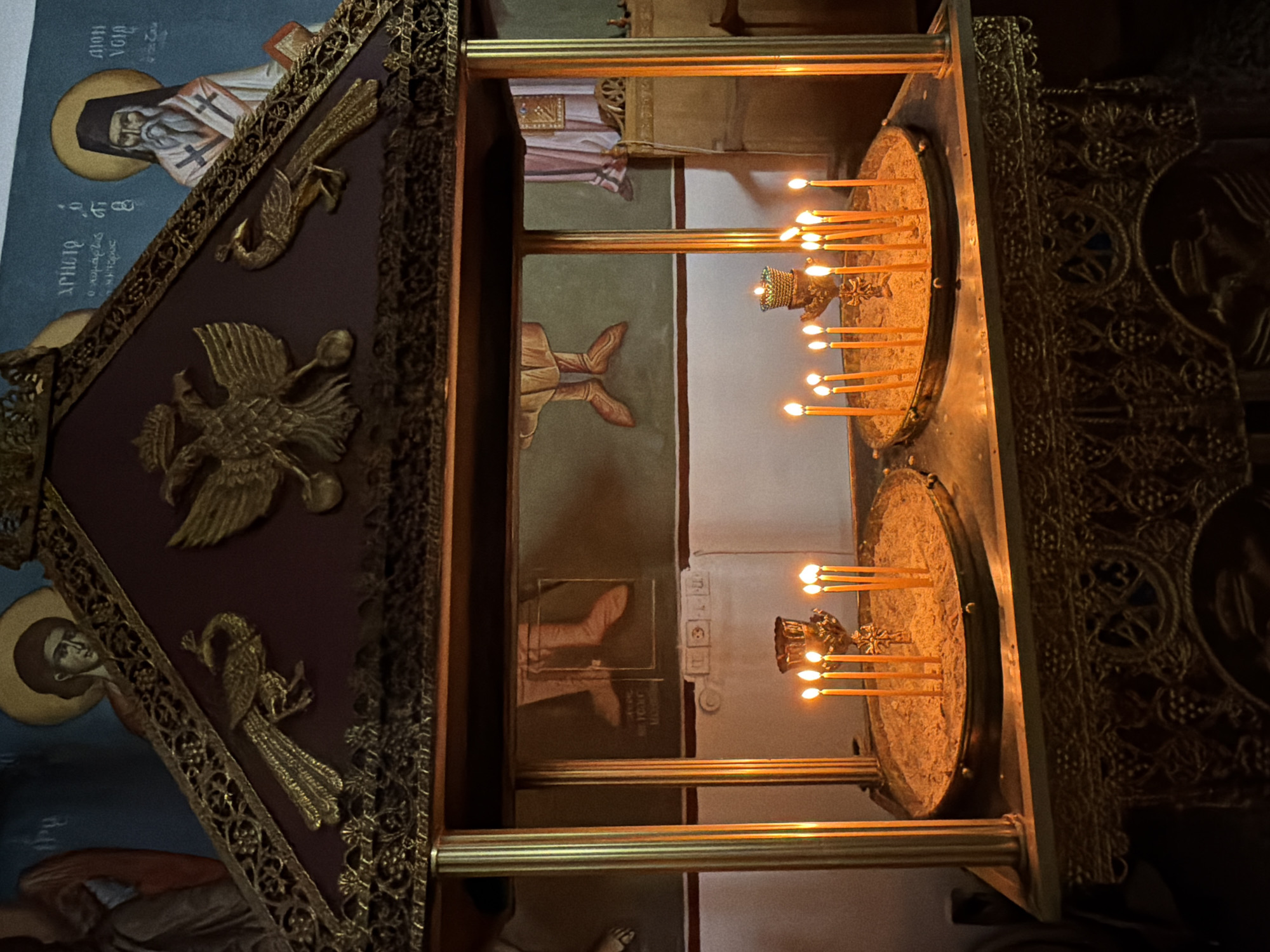 This is a photograph of a candle stand inside a church in the Municipal Unit of Alyzia, Greece. The candle stand is ornate, made of gold-colored metal, and topped with a decorative pediment featuring a double-headed eagle and two peacocks. Below the pediment, there are two circular trays filled with sand, each holding several lit yellow candles. Behind the candle stand, the wall is adorned with religious frescoes depicting figures in halos and robes. The lighting is dim, with the warm glow of the candles illuminating the scene.