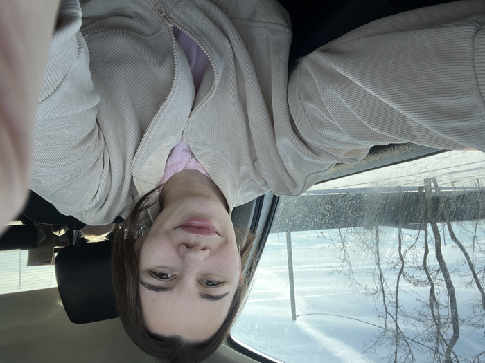 The image depicts a female subject seated inside a vehicle, located in the city of Shchepkin, Russia. The subject has brown hair and a light complexion, looking directly at the camera with a slight smile. She is wearing a light beige, corduroy-textured jacket, partially unzipped to reveal a pink garment underneath, and a thin silver-colored necklace. The subject's right fingers are visible on the far right edge of the frame, indicating a self-portrait. Through the vehicle's driver's side window, which appears dirty or streaked, a winter landscape is visible. This exterior scene includes snow-covered ground, several bare deciduous trees, a dark-colored horizontal fence in the middle ground, and a tall utility pole on the right. The sky is light blue with multiple white contrails.