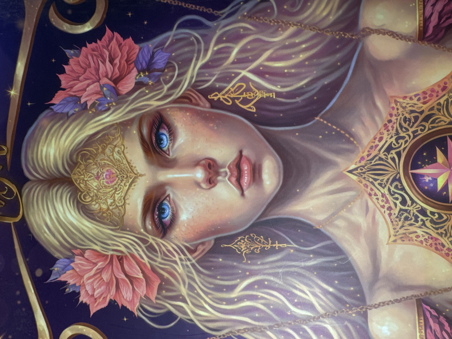 This image depicts a highly stylized, digitally rendered portrait of a female figure, observed in the city of Profesor Ishirkovo, Bulgaria. The subject is shown from the chest up, featuring long, flowing blonde hair parted centrally, adorned with large pink flowers and purple leaf-like accents on either side of her head. A golden, ornate tiara with a pink central gem is positioned on her forehead. Her face presents fair skin with subtle freckles, prominent blue eyes framed by dark eyeliner and long lashes, a straight nose, and full, slightly parted pink lips. She wears elaborate golden, abstract-patterned dangling earrings and a delicate golden chain necklace. On her chest, a significant, ornate golden emblem is displayed, featuring intricate scrollwork and a central circular motif containing a pink multi-pointed star against a dark, starry background. A dark purple/pink draped garment is visible around her shoulders. The background is a dark, cosmic expanse filled with numerous glowing golden specks, and partially visible golden decorative chains and elements, including components resembling the pans of a balance scale on the far left and right. The figure gazes directly forward, establishing direct eye contact.