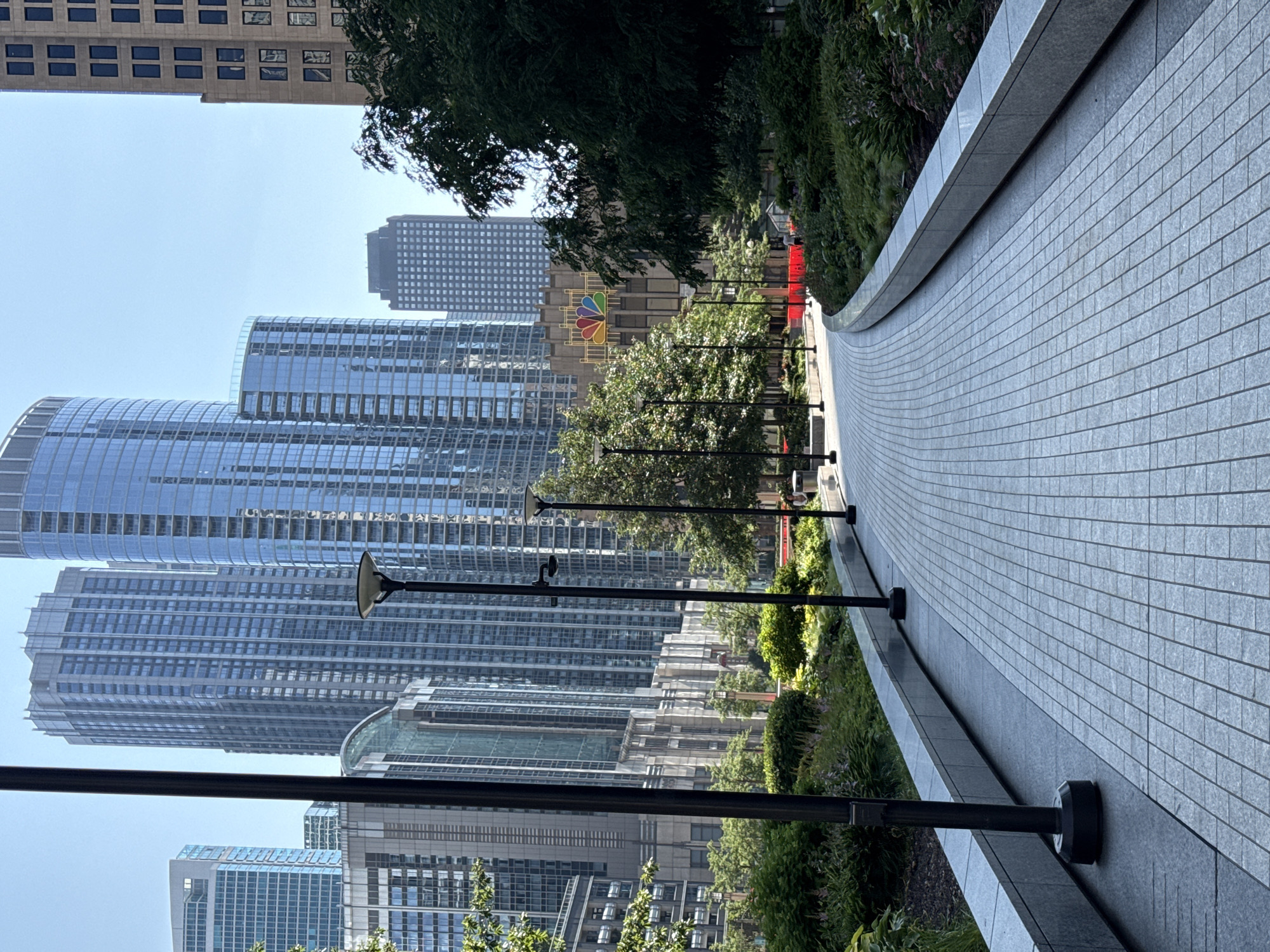 This image depicts a view of the NBC Tower in Chicago, Illinois, USA, on a sunny day. A paved walkway, made of light gray rectangular tiles, curves through a landscaped area with lush green bushes and trees. Several black lampposts are visible along the walkway. In the background, various modern skyscrapers rise into the clear blue sky, some with reflective glass facades. The NBC Tower, a prominent feature, stands out with its distinctive NBC peacock logo prominently displayed. A few people are distantly visible, walking along the path.