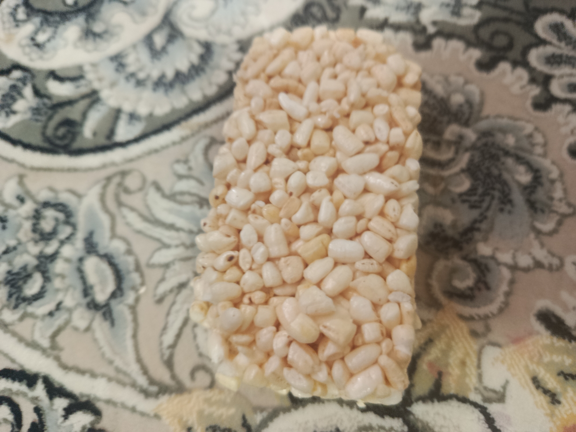A close-up, overhead shot shows a single, rectangular puffed rice snack placed on a patterned textile. The snack is roughly three inches long and one inch wide, composed of tightly packed, irregular pieces of puffed rice. The rice is a pale, creamy yellow, with some pieces exhibiting slightly darker, toasted tips. The textile beneath is a light beige with a repeating floral or paisley pattern in muted blues, grays, and blacks. The texture of the textile appears to be slightly raised or embroidered. The lighting is soft and diffused, creating minimal shadows. The image is sharpest on the snack itself, with the background textile slightly out of focus. The scene is devoid of any human presence or activity, depicting only the static arrangement of the snack and its surface. There are no discernible environmental clues to suggest a specific location within a city.