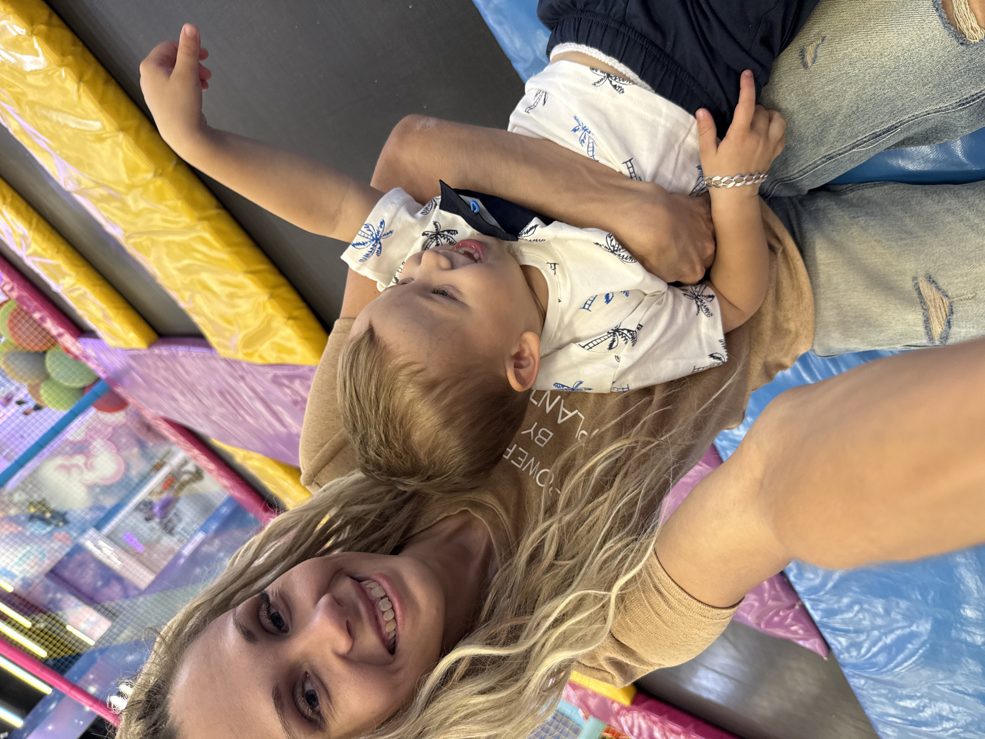 A young blonde woman is taking a selfie with a small child. The woman is wearing a tan t-shirt and ripped jeans. She is smiling at the camera. The child is wearing a white t-shirt with a palm tree pattern and dark shorts. He is looking away from the camera and laughing. They are sitting on a blue mat in what appears to be an indoor playground with yellow and pink inflatable structures in the background. The location is not identifiable as Zhukovo, Russia based on the visual information provided.