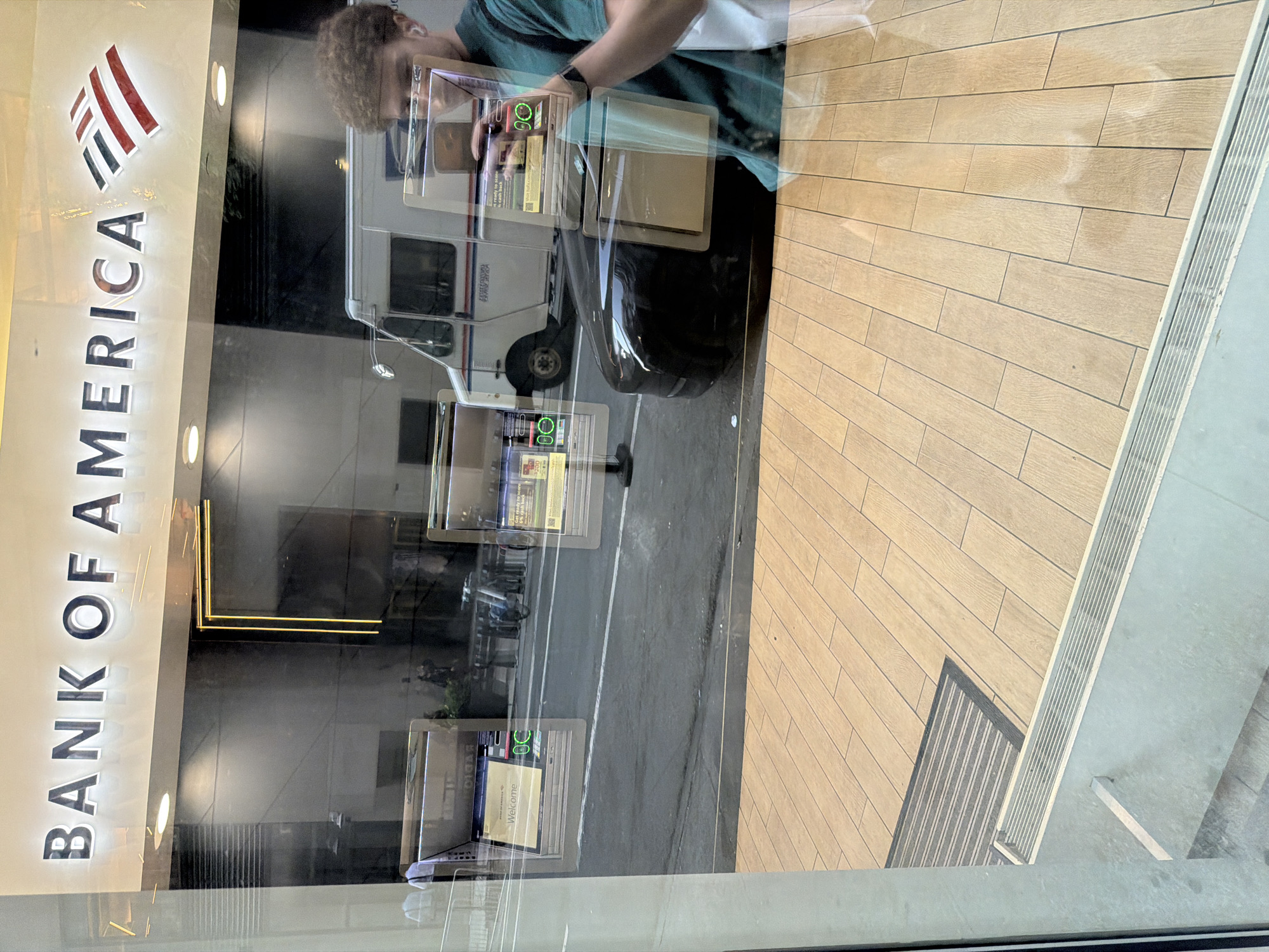 The image shows a Bank of America branch in New York City. Reflected in the window is a person holding a phone and a white USPS mail truck is parked outside the branch. The interior of the branch has automatic teller machines. The floor inside the branch is made of wood and a floor mat is located near the entrance.