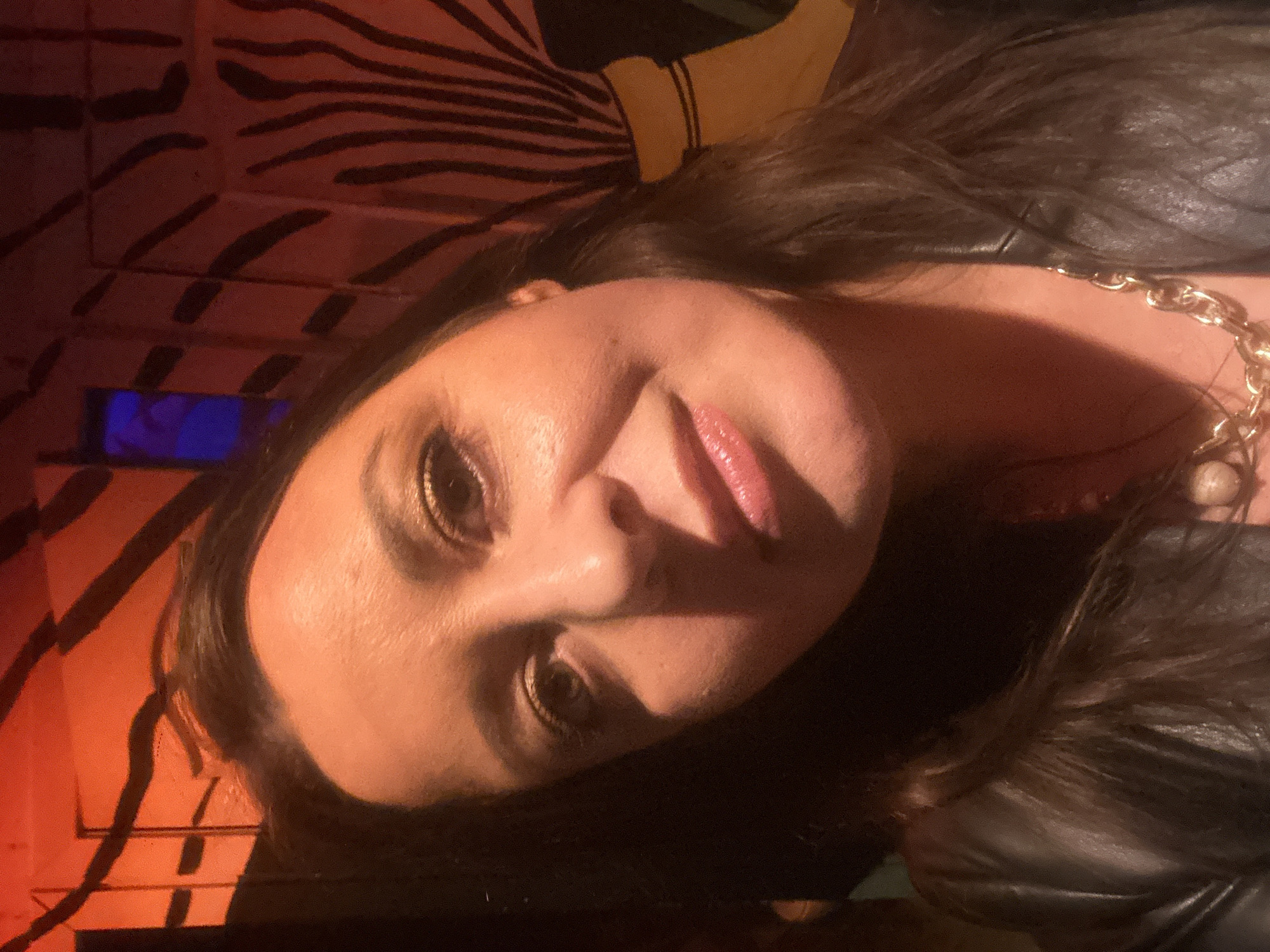 A woman with dark hair is shown from a tilted head-and-shoulders perspective, looking at the camera with a slight smile. She is wearing a dark leather jacket and a chunky gold chain necklace with a pearl. Her eyes are made up with gold eyeshadow, and her lips have a glossy pink shade. The background consists of a wall featuring a zebra-stripe pattern in tones of orange and black. A blue shape appears to be part of the decor behind her. There's no visible evidence to confirm the location is Riga, Latvia, or any specific city. No actions or interactions are taking place in the frame. The environment is defined by the indoor lighting and the patterned background.