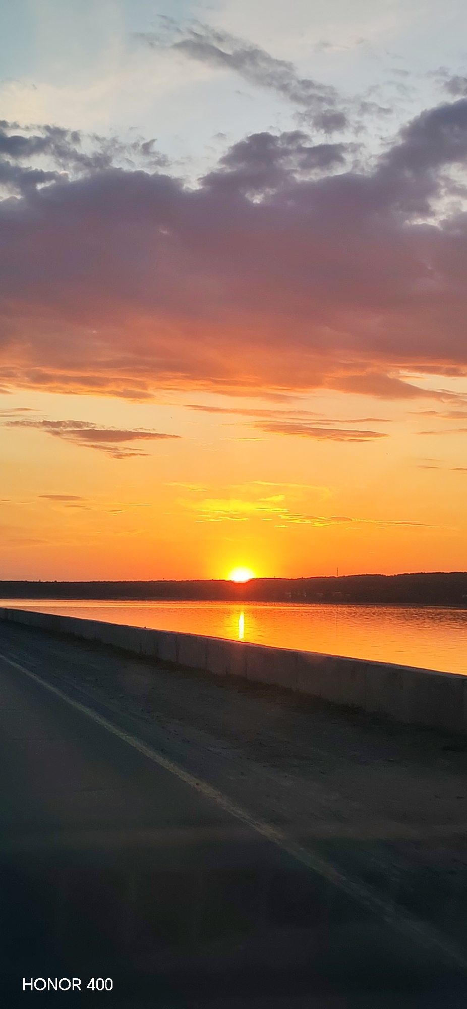 The image shows a sunset over the Kama River in the city of Chaikovsky, Perm Krai, Russia. The sun is setting on the horizon, creating a bright orange glow that is reflected in the water. There is a concrete embankment in the foreground and a road. The sky is filled with colorful clouds, ranging from shades of orange and pink to gray and blue. The text "HONOR 400" is in the bottom left corner of the image.