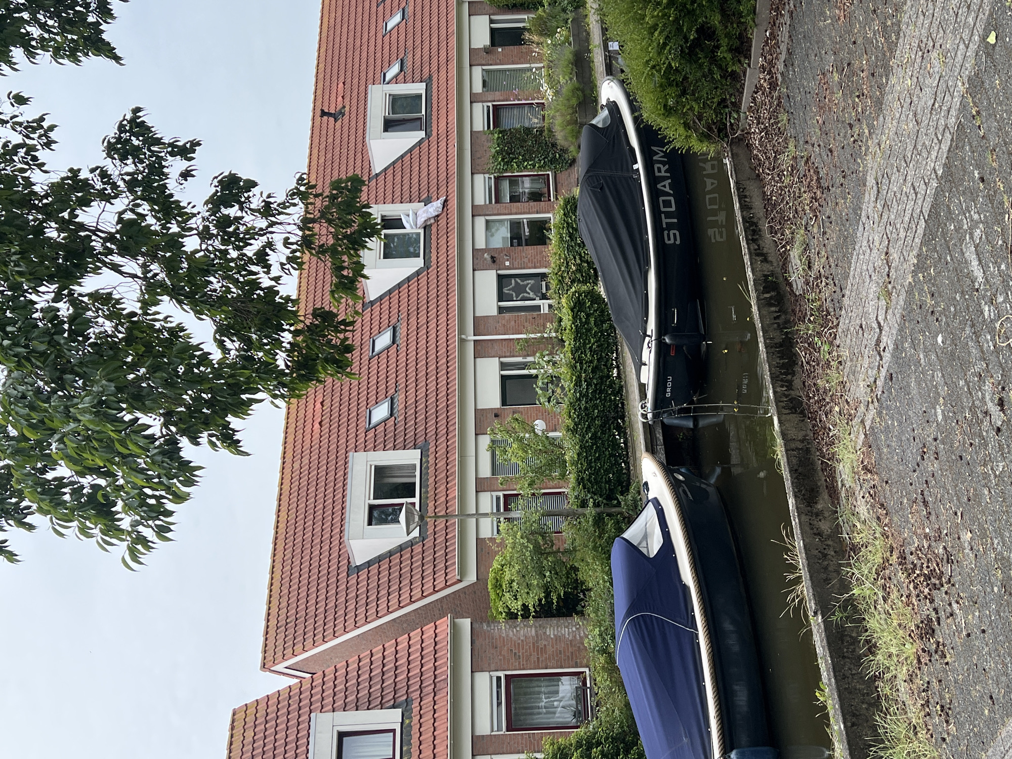 This is a photograph taken in the city of Grou, Netherlands, capturing a canal-side scene with residential buildings and boats. The focus is on the waterway, with two boats moored along its edge. The boat closest to the viewer is covered with a dark blue tarp, and its hull is partially visible, displaying a white trim. Further down the canal, a second boat is covered with a black tarp. The name "STOARM" is clearly visible on the side of this second boat.
Behind the canal, a row of brick houses with red-tiled roofs stands. These houses feature dormer windows and standard windows, some of which have blinds. A lush green hedge runs along the front of the properties, bordering the canal. Trees with dark green leaves are visible in the foreground, partially obscuring the upper parts of the houses and the sky. The sky is overcast, suggesting a cloudy day. The ground in front of the canal is paved with interlocking bricks and has some loose debris scattered on it. A lamppost is positioned between the houses and the canal.