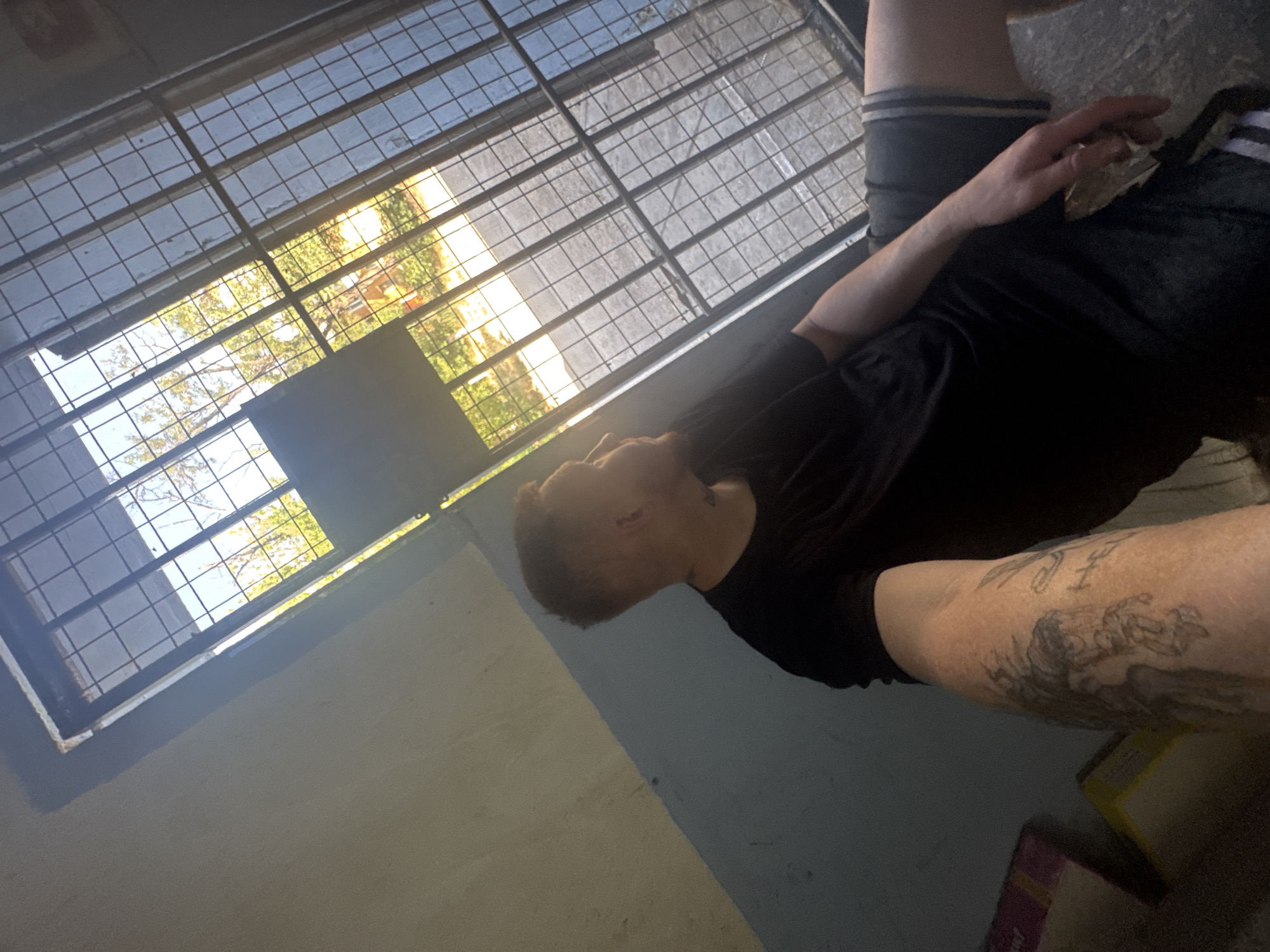 A medium shot captures a fair-skinned male with short, light brown hair and a beard, looking away from the camera. He is wearing a black t-shirt and dark grey shorts with white stripes. The man has tattoos on his left arm and a tattoo on his neck. He is seated in an indoor space with a concrete floor and a wall that is half light grey and half light blue. A large window with a metal grate covers the upper portion of the scene, revealing an outdoor view of trees and sunlight. The perspective is from a low angle, looking upwards.