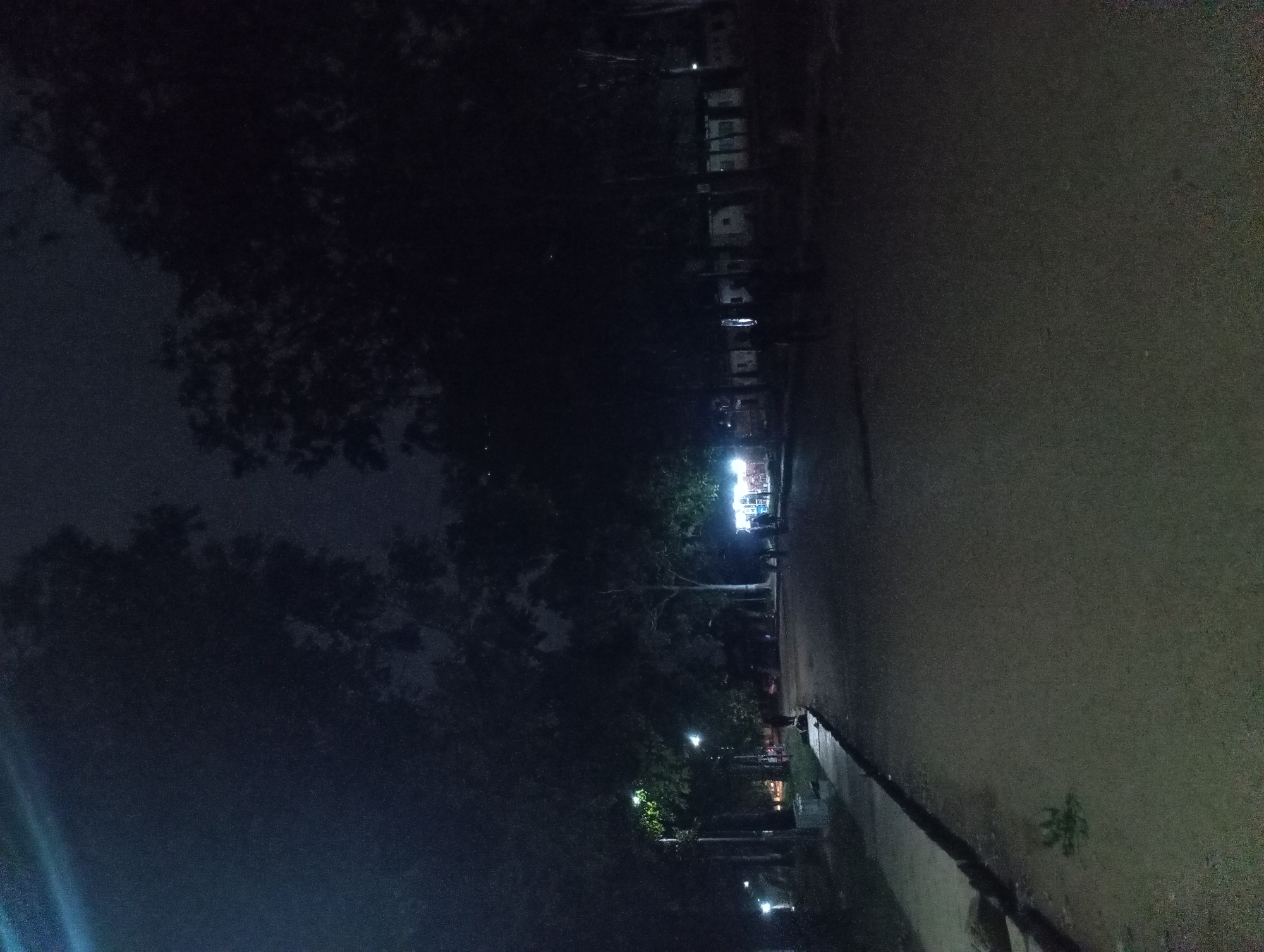In the city of Unknown, Unknown, a dimly lit scene unfolds at night, possibly in a park or a quiet street. A paved pathway curves through the foreground, flanked by a raised concrete curb and illuminated by scattered streetlights. To the left, trees with dark foliage create a dense canopy, with a few streetlights casting pools of light on the ground. In the mid-ground, several figures are visible, some walking and others gathered near a brightly lit stall or shop. The area is surrounded by more trees and some low-lying structures that are partially obscured by darkness. The overall atmosphere is serene and somewhat mysterious due to the low light conditions and the presence of scattered artificial illumination.