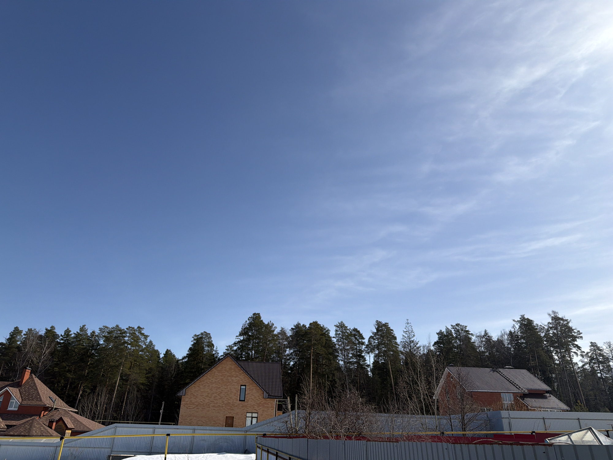 The image depicts a daytime scene in Chaikovsky, Russia, under a clear blue sky with wispy clouds. In the foreground, a grey metal fence with red trim is partially visible. Behind the fence, a sparse layer of snow is present. Two brick houses with dark, gabled roofs are visible among tall evergreen trees. The house on the left is a lighter orange brick, while the one on the right is a darker red brick. The houses appear to be situated within a residential area surrounded by a dense forest. There are no people visible in the image.