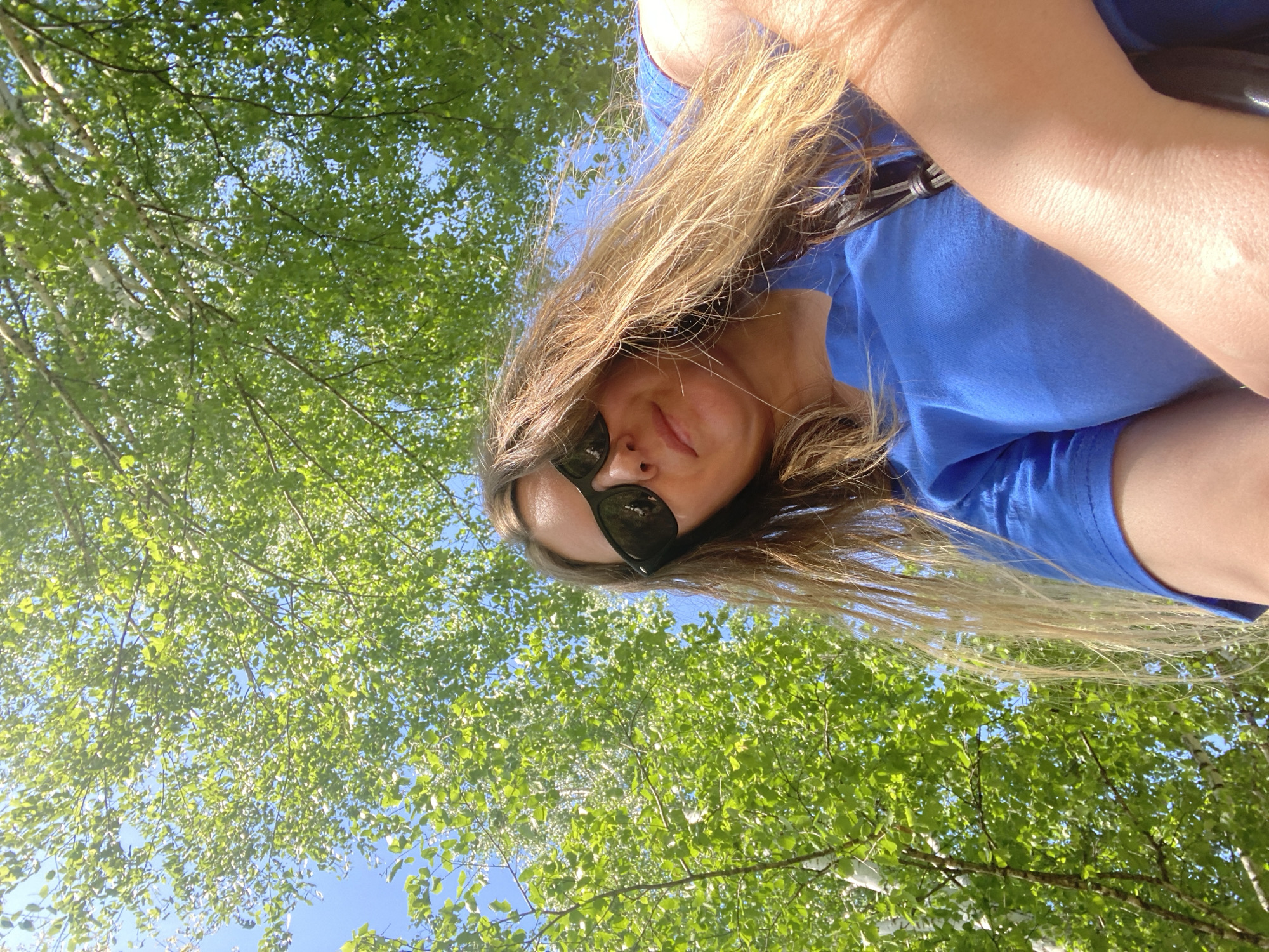 A low-angle shot looking up at a woman wearing sunglasses and a blue t-shirt, with a background of green leaves and a partially visible blue sky. The location is identifiable as Noviy, Russia based on visual characteristics such as tree species and architectural styles common to the region.