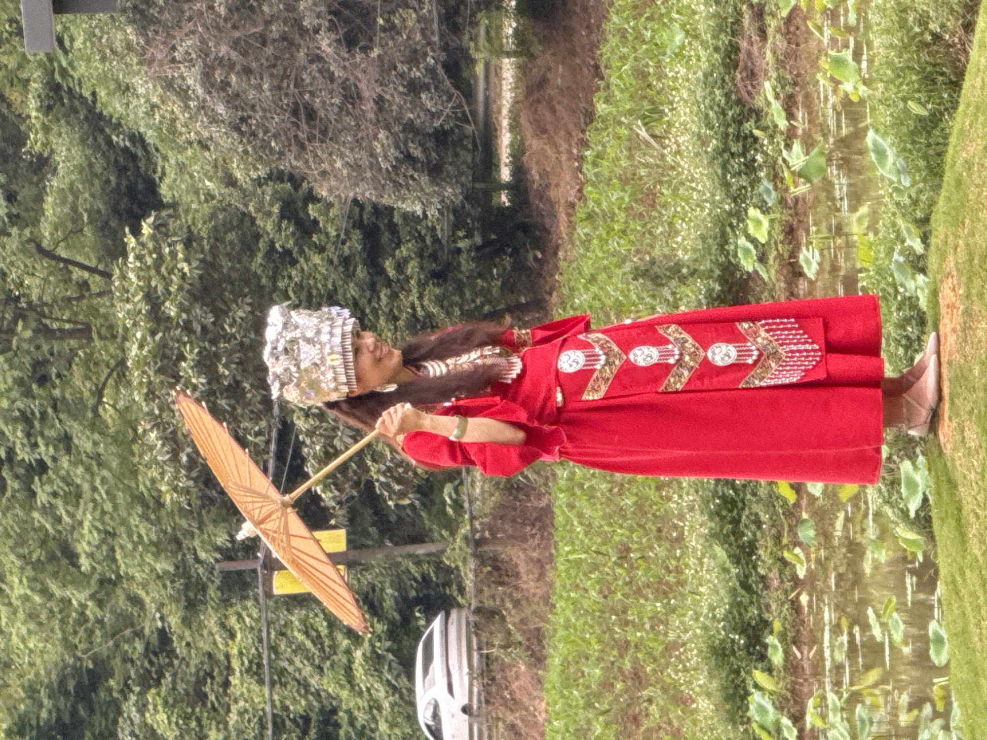 A woman in traditional red dress with silver headwear is standing next to a body of water in Guilin, China. She is holding an open, light-colored umbrella. The background includes trees and foliage. There is a car and a utility pole visible in the distance. The body of water contains scattered aquatic plants.