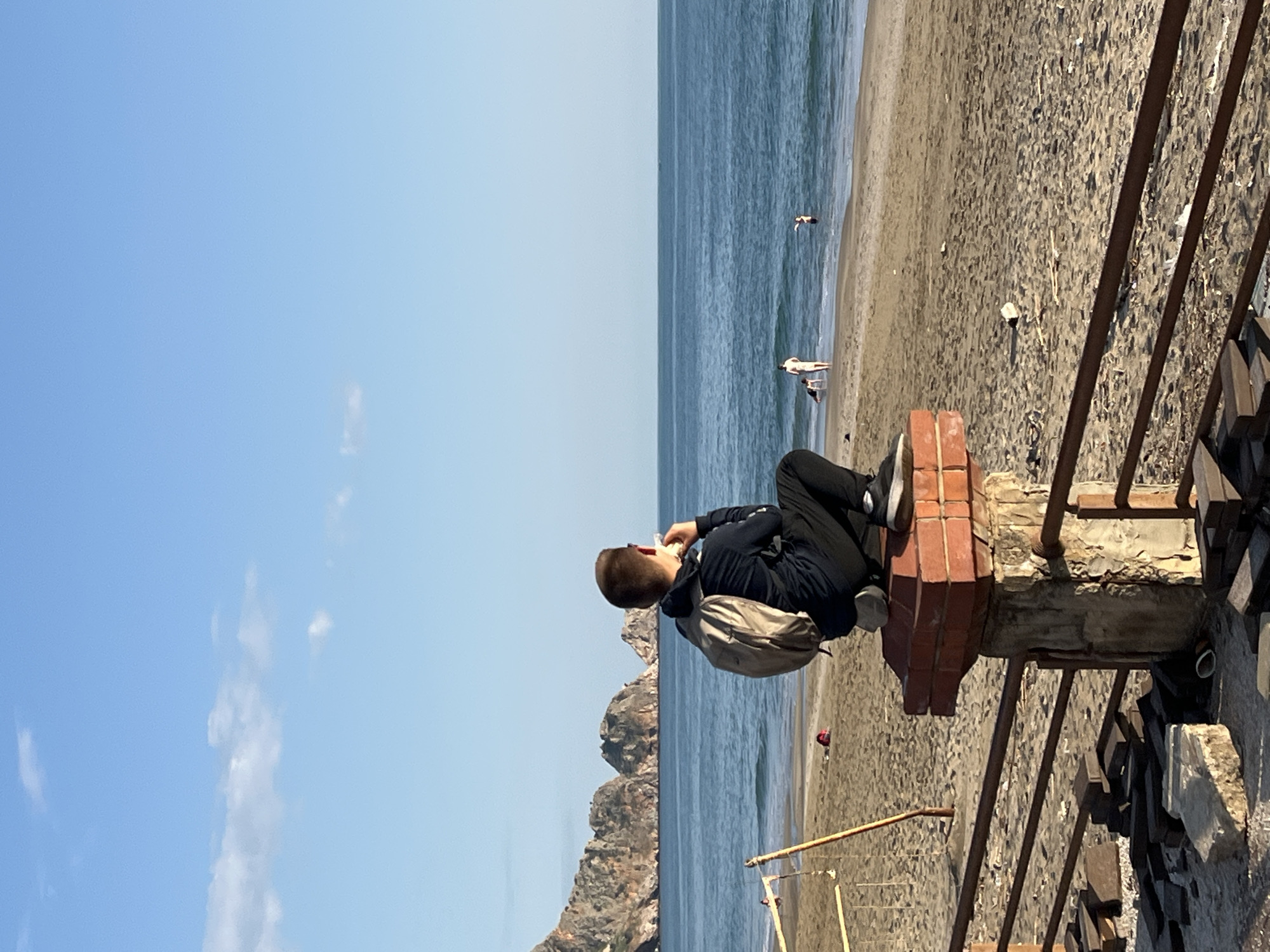 A person sits on a brick and concrete structure overlooking a beach and the ocean in what appears to be Alanya, Turkey. The person is wearing dark clothing, a backpack, and white sneakers. The person is turned towards the ocean, possibly eating something. The beach has several people on it, and mountains are visible in the distance. A metal railing is in the foreground. The sky is blue with a few clouds.