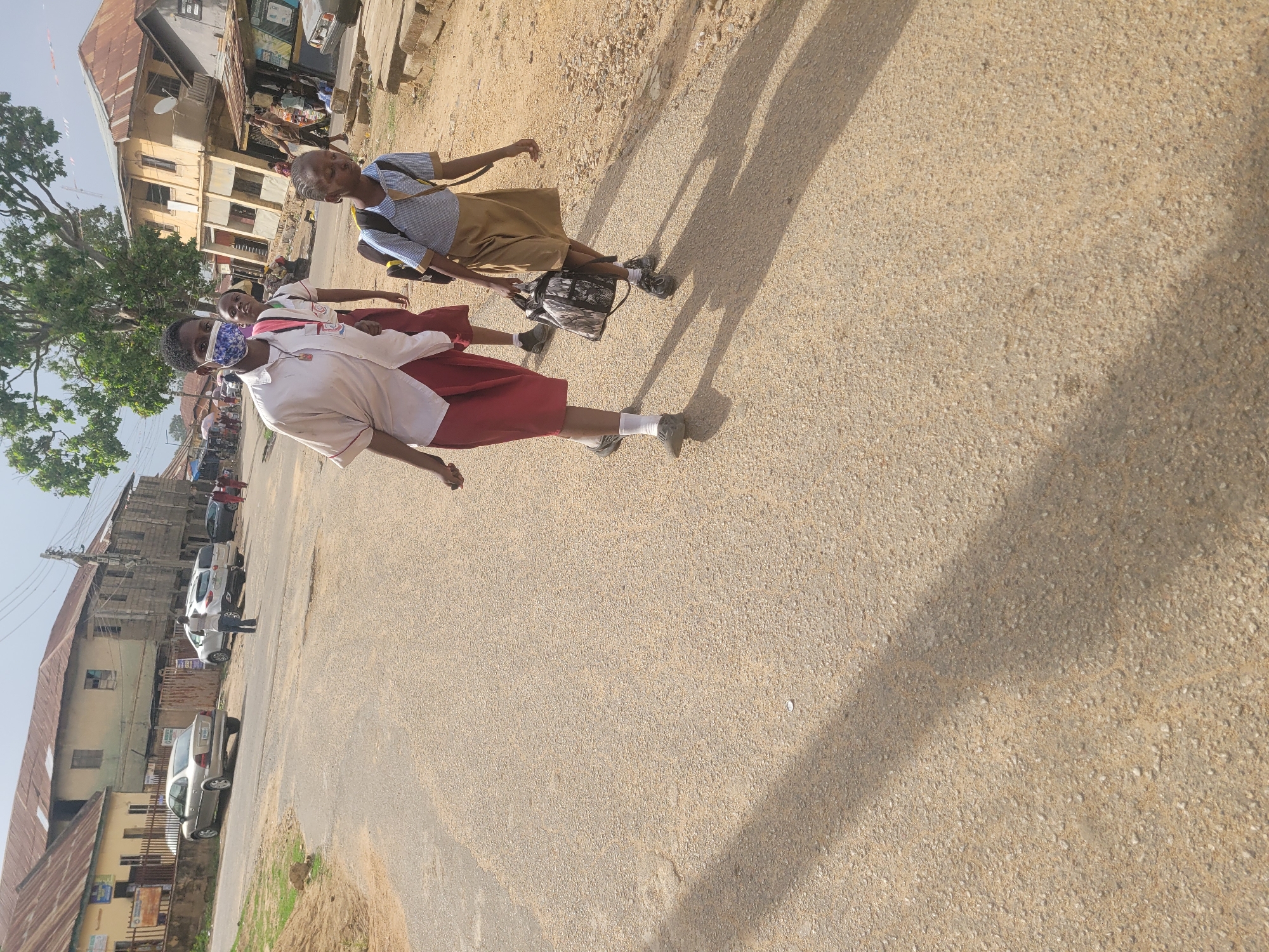 The image shows two children walking on a paved street in Jos, Nigeria. The child in the foreground wears a white shirt, red skirt, and a patterned face mask. The other child is behind them, wearing a blue shirt and tan skirt, carrying a backpack. Buildings are visible in the background, along with parked cars. The scene is illuminated by sunlight.