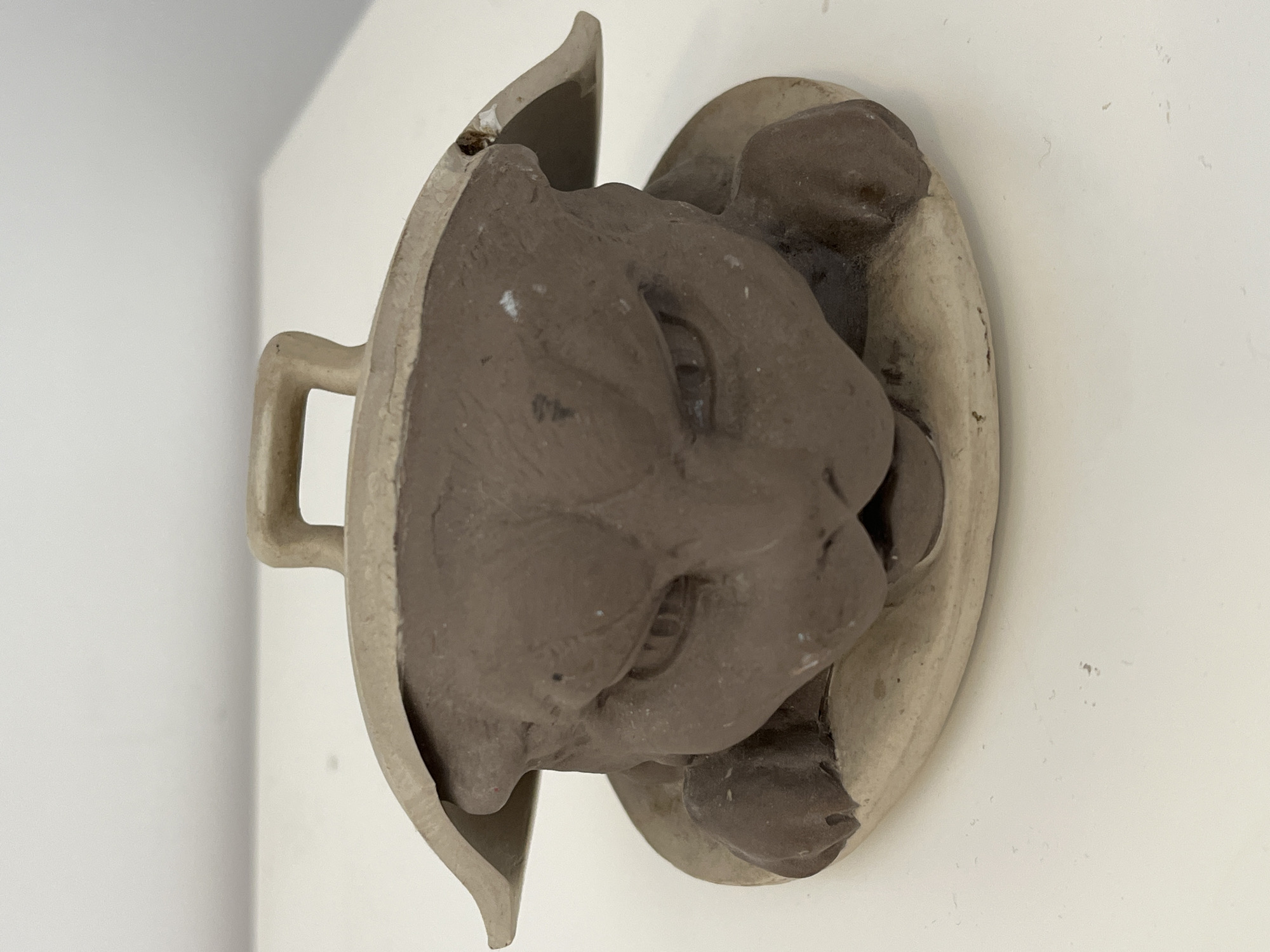 A close-up shot shows a broken ceramic object mounted on a white wall. The object is mostly light gray, with a darker gray, sculpted lion's head protruding from within. The ceramic is cracked, and a piece is visibly broken off at the top. A handle is present on the left side of the object. The background is a plain white wall. There is no evidence suggesting the location is Chambœuf, France.