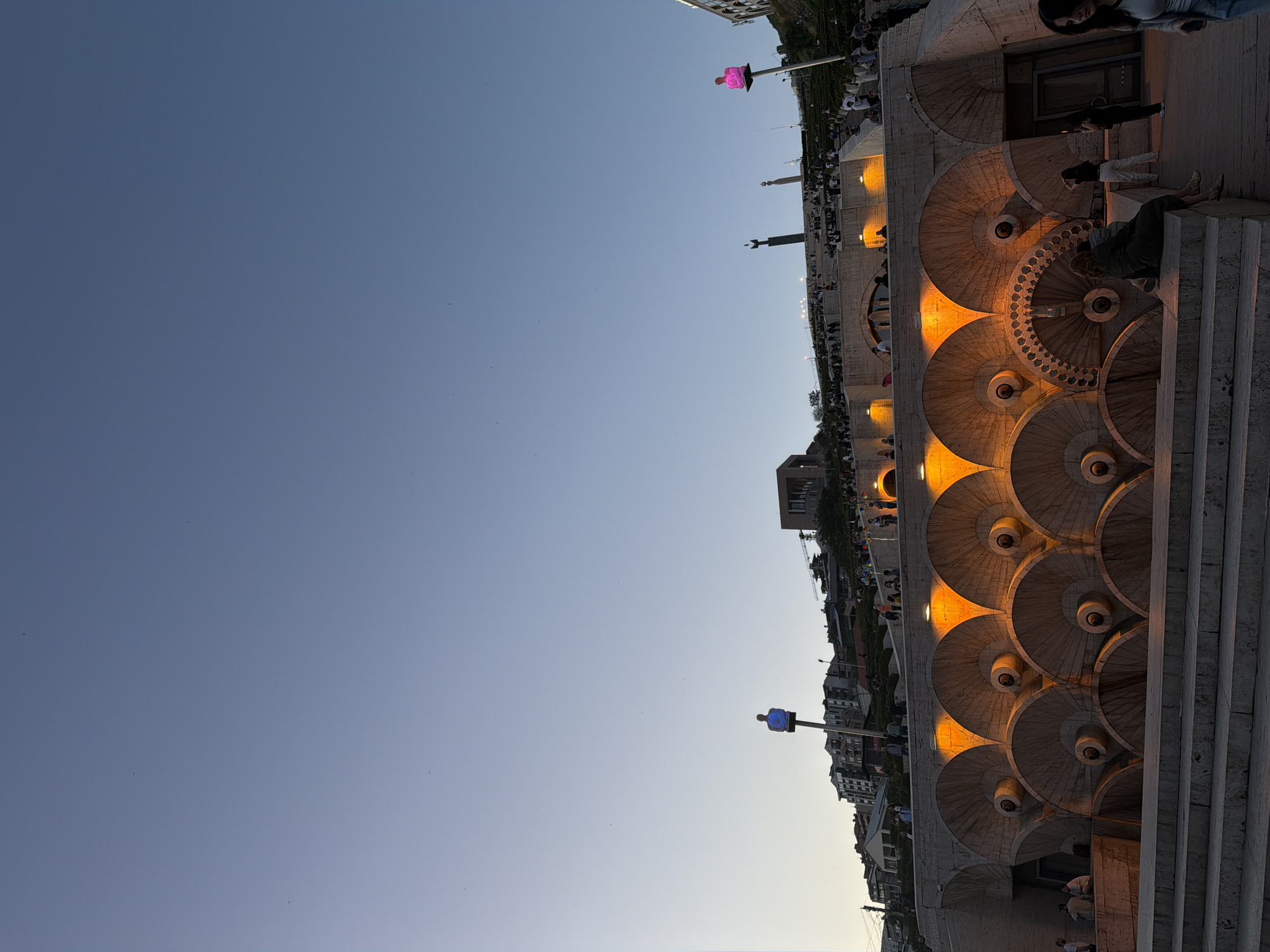 The image shows a section of the Yerevan Cascade in Armenia. The Cascade is a monumental staircase with fountains and sculptures, ascending a hillside. The foreground features a stone staircase, with semicircular water features cascading down each level. These features are illuminated with orange light. In the background, there are buildings and monuments, including one with a bright pink light on top. People are visible walking and sitting near the staircase. The sky is clear and blue.
