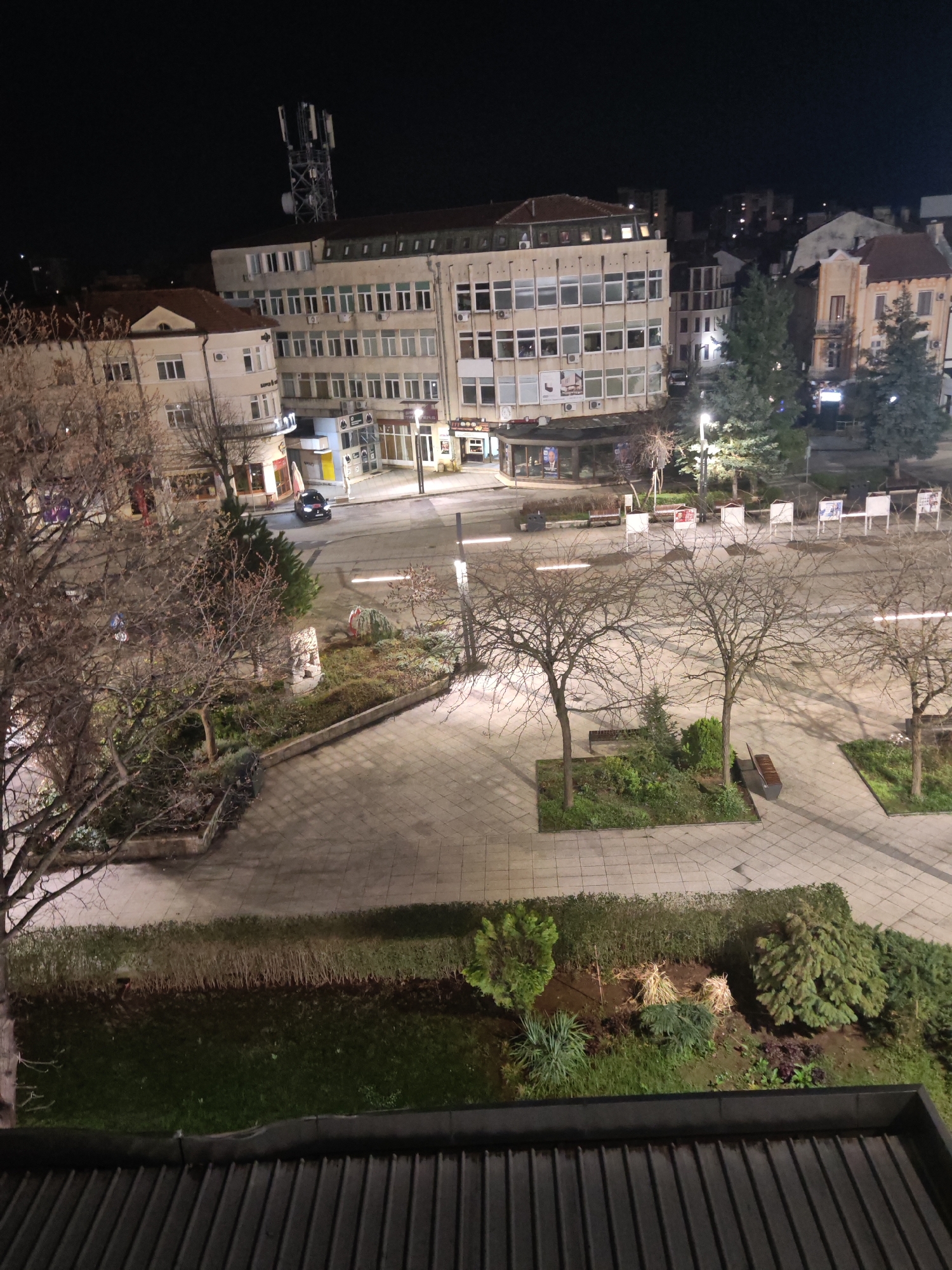 The image is a nighttime view of a town square in Vratsa, Bulgaria, taken from an elevated position, likely a building. The square is paved with large rectangular tiles and contains landscaped areas with grass, bushes, and trees, mostly bare of leaves. Benches are situated near some of the trees.
Several buildings surround the square, including a multi-story office building with a communication tower on its roof. Shops and businesses occupy the ground floors of these buildings. Streetlights illuminate the square and surrounding streets.
A car is visible on the road passing through the square. Signboards or displays are arranged in a row along one side of the square. The foreground of the image includes a view of a rooftop with dark corrugated panels.
