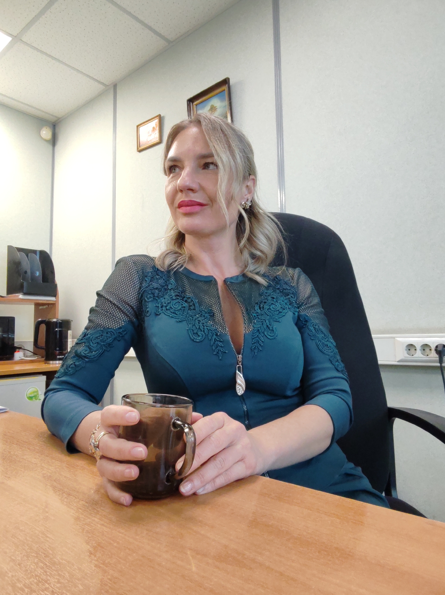 A blonde woman with reddish-pink lipstick sits in a chair at a wooden desk, holding a dark glass mug filled with a brown liquid. She is wearing a dark teal dress with sheer, patterned netting on the sleeves and chest. The dress has a zippered front with a silver pull tab. She has blonde hair styled in loose waves and is wearing earrings and a ring on her left hand. The background shows an office environment with a white tiled ceiling, light-colored walls, and framed pictures hanging on the wall. To the left of the woman, there is a shelf with a black document holder and what appears to be a black electric kettle. To the right, a white electrical outlet strip is visible on the wall. The woman is looking away from the camera towards the left.
