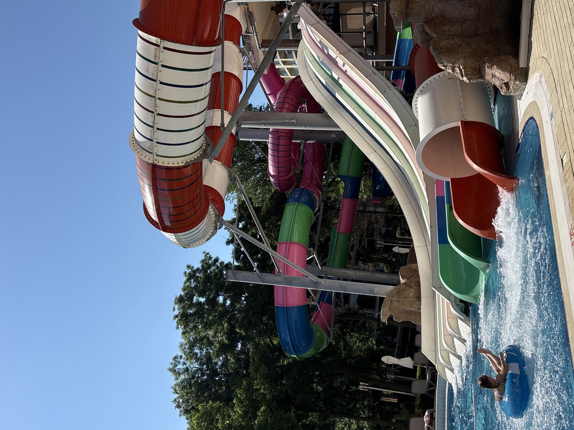 This image captures a vibrant and active scene at a water park, likely located in Nesebar, Bulgaria. The central focus is a complex network of colorful water slides, set against a clear blue sky on a bright, sunny day.
Multiple water slides are visible, varying in design and color. On the left, several large, enclosed tube slides feature a striking red and white striped pattern, with thin horizontal bands of various colors accenting the segments. Towards the center, other slides include solid pink tubes, some with internal stripes, as well as green and blue segmented tubes. On the right, an open, multi-lane slide displays pastel-colored stripes (pink, green, light blue, beige, and white), resembling a rainbow flume, leading into the main pool. Another red, open spiral slide is also prominent.
In the bottom right of the frame, a child or young person is seen riding a blue inflatable tube, emerging from one of the slides and splashing into the landing pool. The tube has "AQUA RIO" printed on its side. The pool water is clear and blue, with significant splashing around the slide exits. The tiled edge of the pool is visible, and a red marker with "1.2 M" can be seen on the rim, likely indicating water depth.
The entire slide structure is supported by a robust metal framework. Lush green trees are visible in the background, contrasting with the bright colors of the slides and the sky. Artificial rock formations are integrated into the landscape around the pool and slides. The clear sky and bright lighting indicate it is daytime, probably midday or early afternoon, suggesting ideal weather for water park activities.