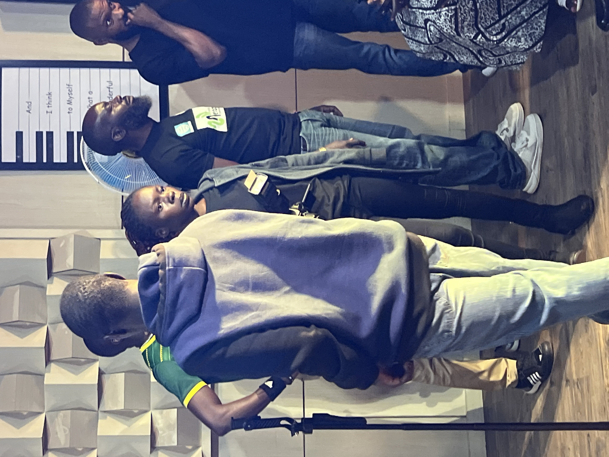In what appears to be an indoor setting, possibly a studio or event space, several individuals are captured in a candid moment. The backdrop features a textured wall with a geometric pattern in shades of beige and grey. To the right, a framed piece of art with text and stylized musical notes is visible. A fan is situated between the artwork and a group of people.
In the foreground, the back of a person wearing a purple hooded sweatshirt and light blue jeans is prominent. To their left, partially obscured, is another individual in a green shirt with yellow trim. Further to the right, a woman with dark braided hair is looking towards the left. Next to her, a man with a beard is looking to the right. Another man, wearing a black t-shirt and dark jeans, stands to the far right.
The individuals are standing on a wooden floor. The lighting in the scene is a mix of artificial light, casting shadows and highlighting certain areas. The overall atmosphere suggests a gathering, perhaps during a break or while waiting for an event to begin. The location in the city of Jos, Nigeria, is not evident from the visual cues in the image.