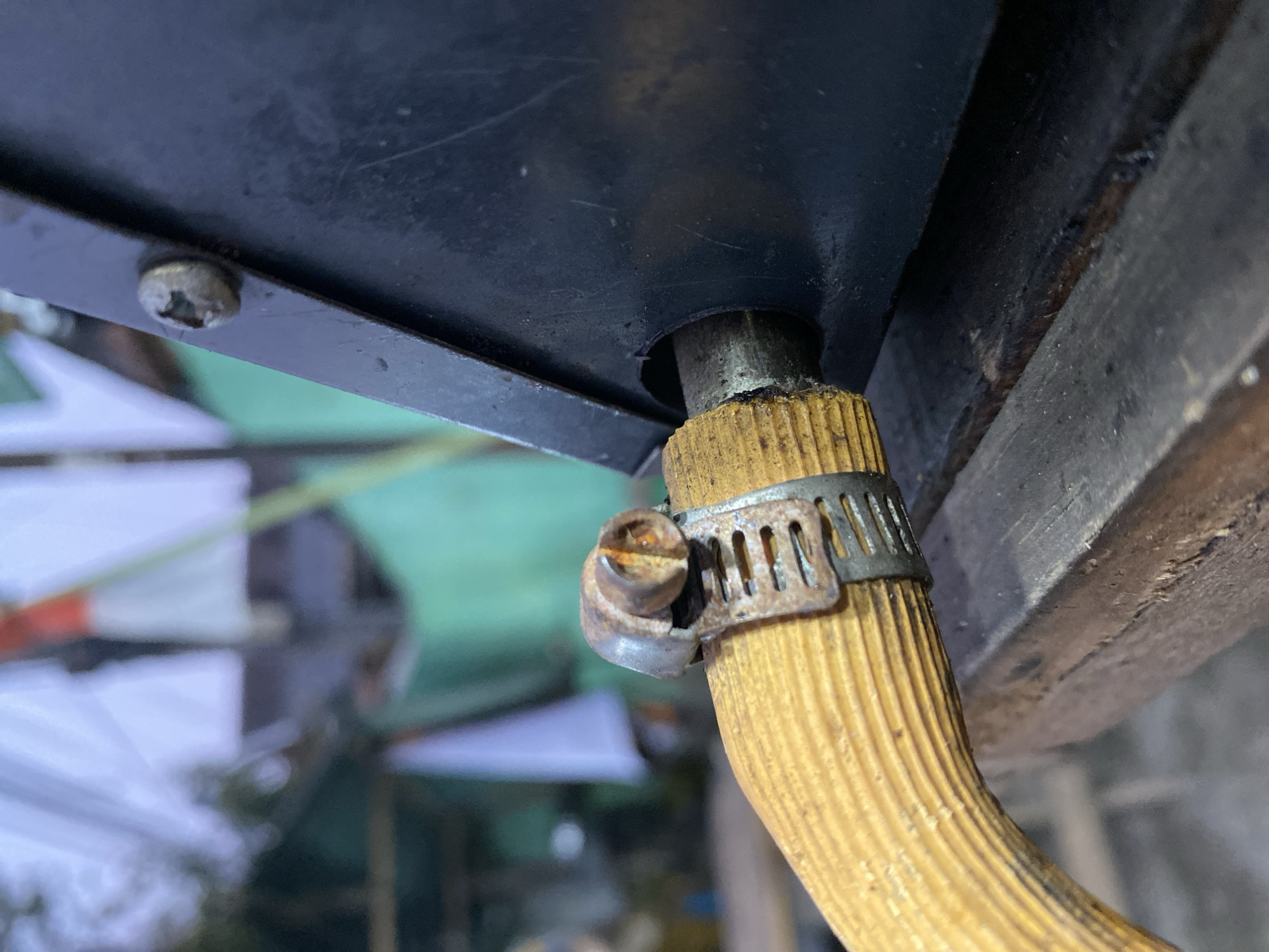 The image shows a close-up of a yellow hose secured by a rusted metal clamp to a black apparatus. The hose has a ribbed texture. The clamp is tightened with a screw mechanism. In the background, out of focus, are what appear to be white and green tarpaulins, and possibly flags, suggesting an outdoor setting. A single screw is visible on the black apparatus, likely fastening it to something. The overall impression is of a functional, but weathered, setup. The scene is likely located in Kasihan, Indonesia, based on the provided information.