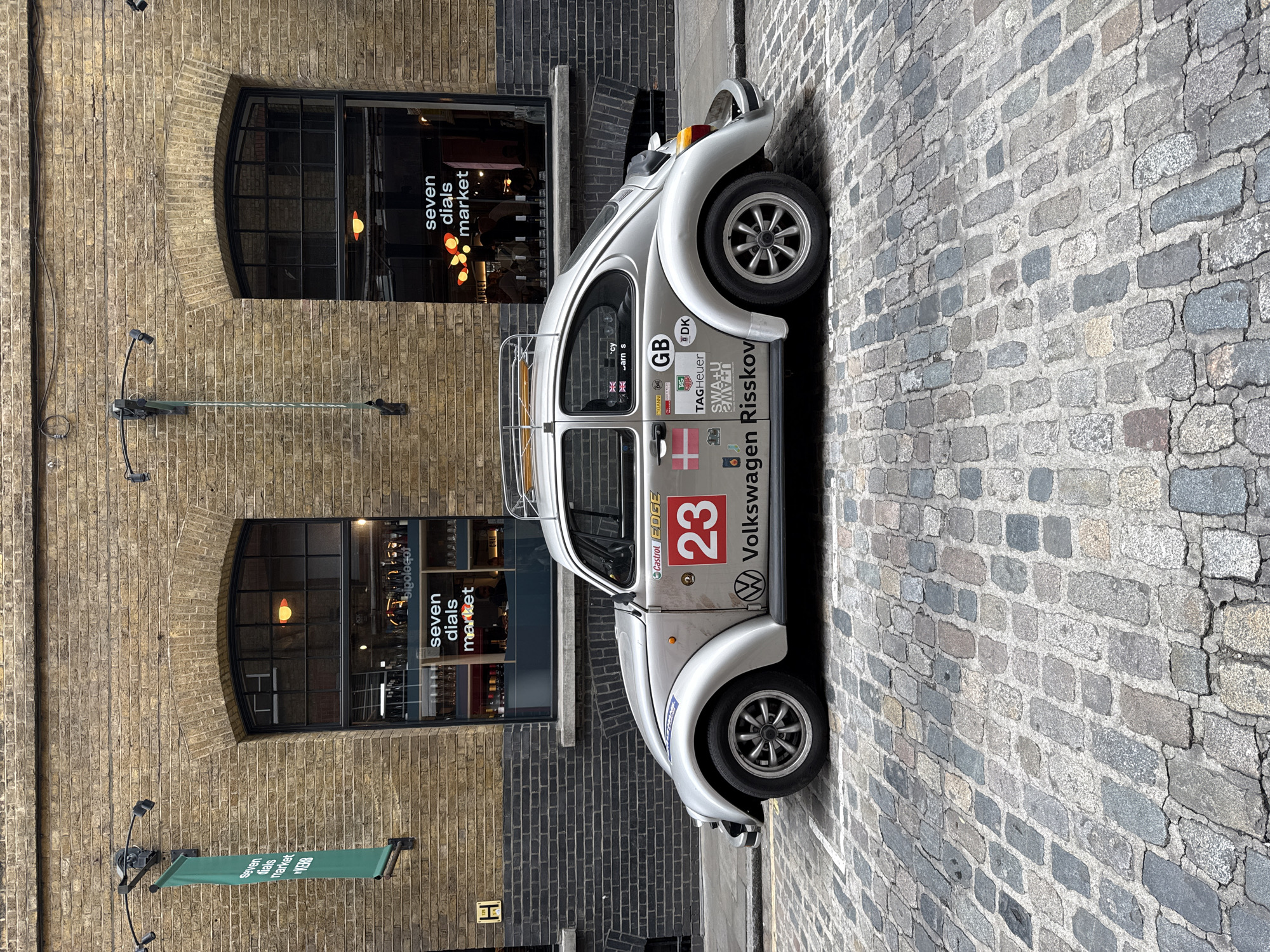 A silver Volkswagen Beetle is parked on a cobblestone street in London, UK, next to a brick building. The car is facing right. It has stickers and the number "23" prominently displayed. "Volkswagen Risskov" is also written on the car. The building behind the car has two arched windows, each displaying "seven dials market" text. A sign displaying the same text hangs beneath.