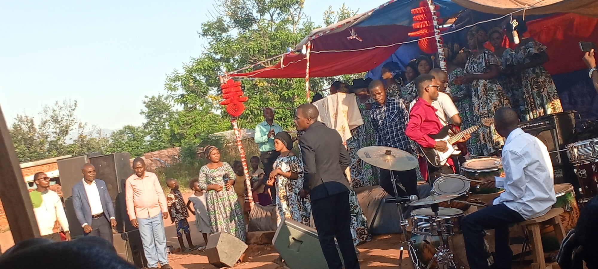 The image shows an outdoor gathering, likely in the city of Kalemie, Democratic Republic of the Congo. A group of people, including a choir in patterned dresses, are on a raised platform covered by a red and blue canopy decorated with red lanterns. A band is performing, with a guitarist in a red shirt and a drummer visible. Speakers and musical equipment are present. Several individuals, including men in suits and casual attire, stand in front of the platform. Children are also present. The backdrop includes trees and what appears to be a hillside.