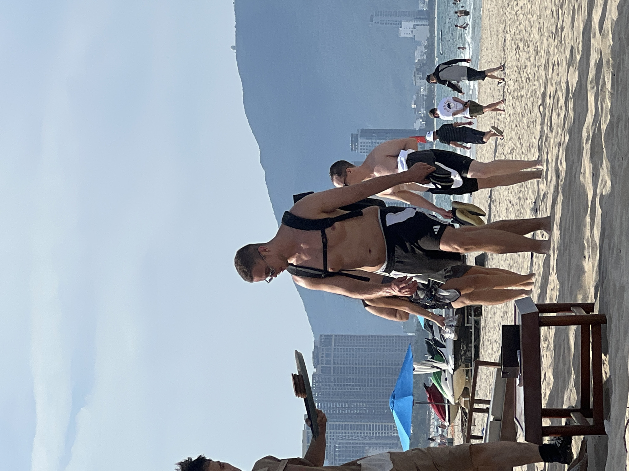 The image captures a sunny day at My Khe Beach in Đà Nẵng, Vietnam. Several individuals are visible on the sandy shore. In the foreground, a person dressed in tan shorts and a white garment is partially visible, holding a tray with items. To their right, a man with a backpack and sunglasses is walking, wearing black shorts. Behind them, other beachgoers are seen, some walking and others in the water. Jet skis are parked on the beach near a blue umbrella. In the background, the cityscape of Đà Nẵng with its modern buildings and the Sơn Trà Peninsula with its characteristic spherical structure are discernible. The sky is clear with some light clouds.